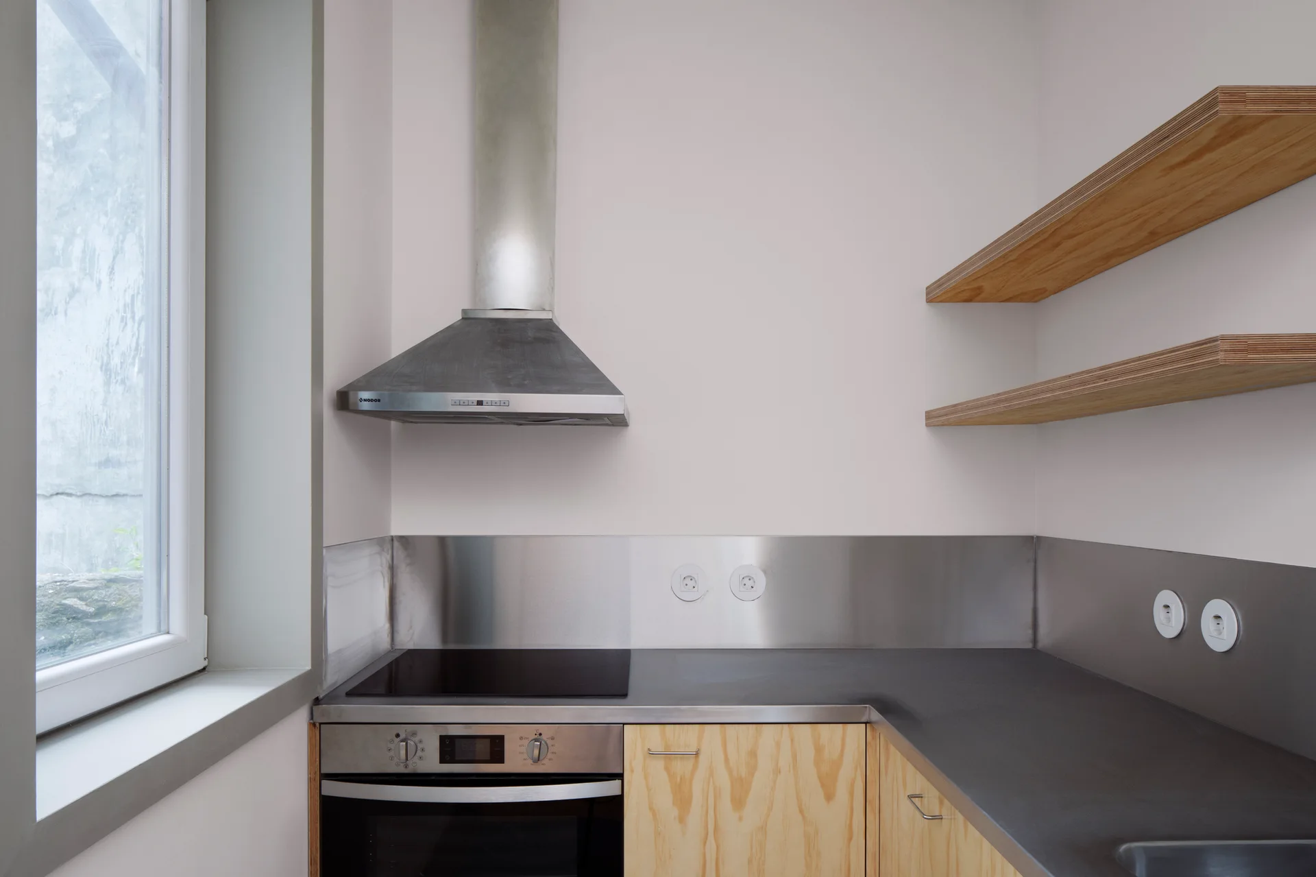 A minimalist kitchen corner featuring plywood cabinets, a stainless steel backsplash and range hood, and floating wooden shelves on white walls.