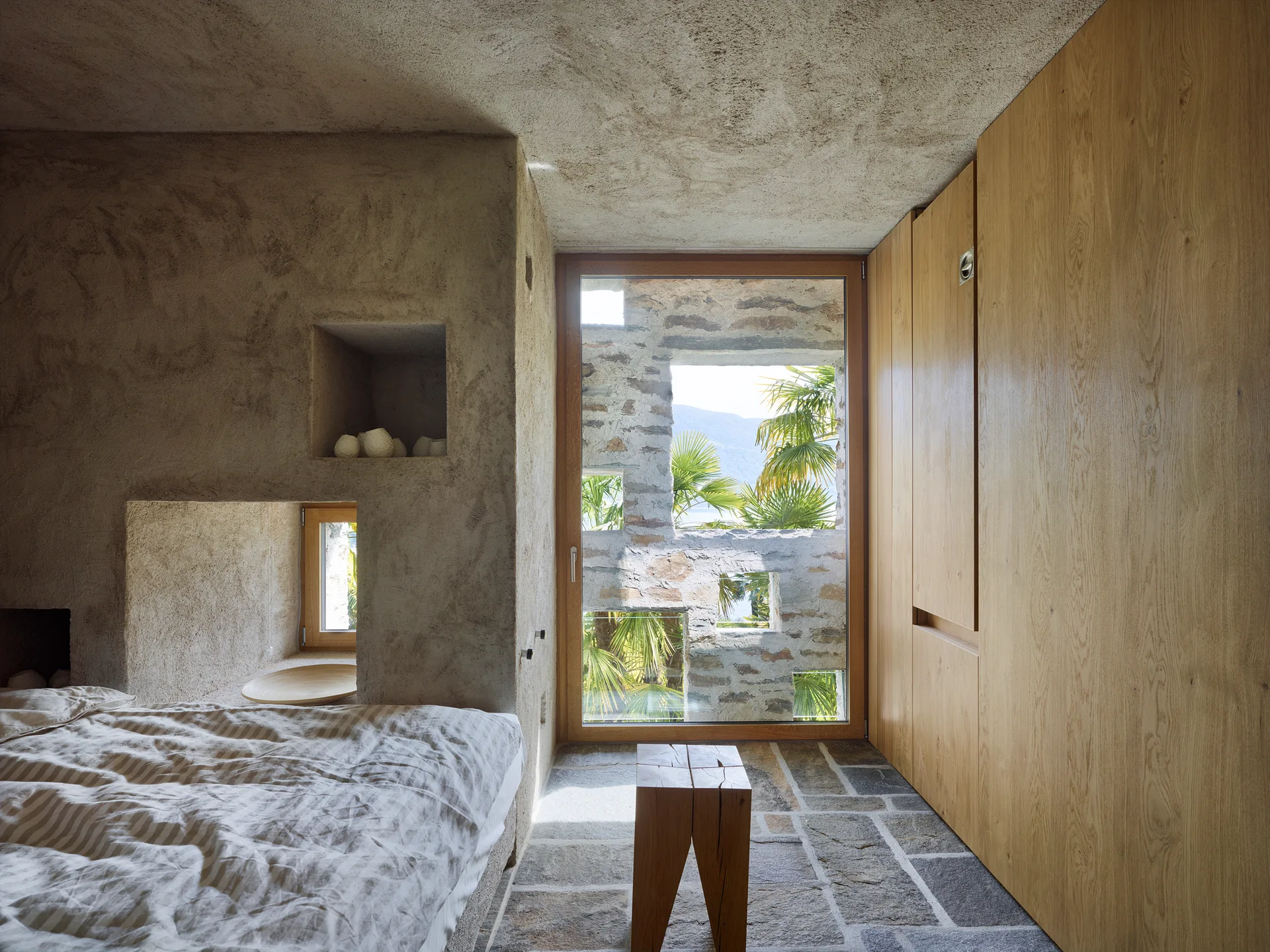A rustic bedroom features a bed with striped bedding, textured plaster walls with built-in niches, and an irregular stone tile floor.