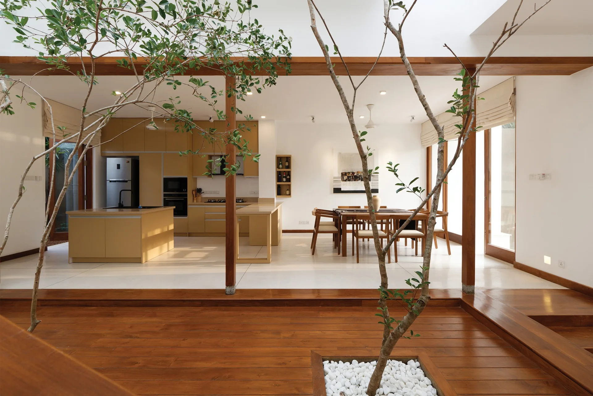 An open-plan kitchen and dining area features tan cabinetry and a wooden dining set, framed by indoor trees growing from a raised wooden platform.