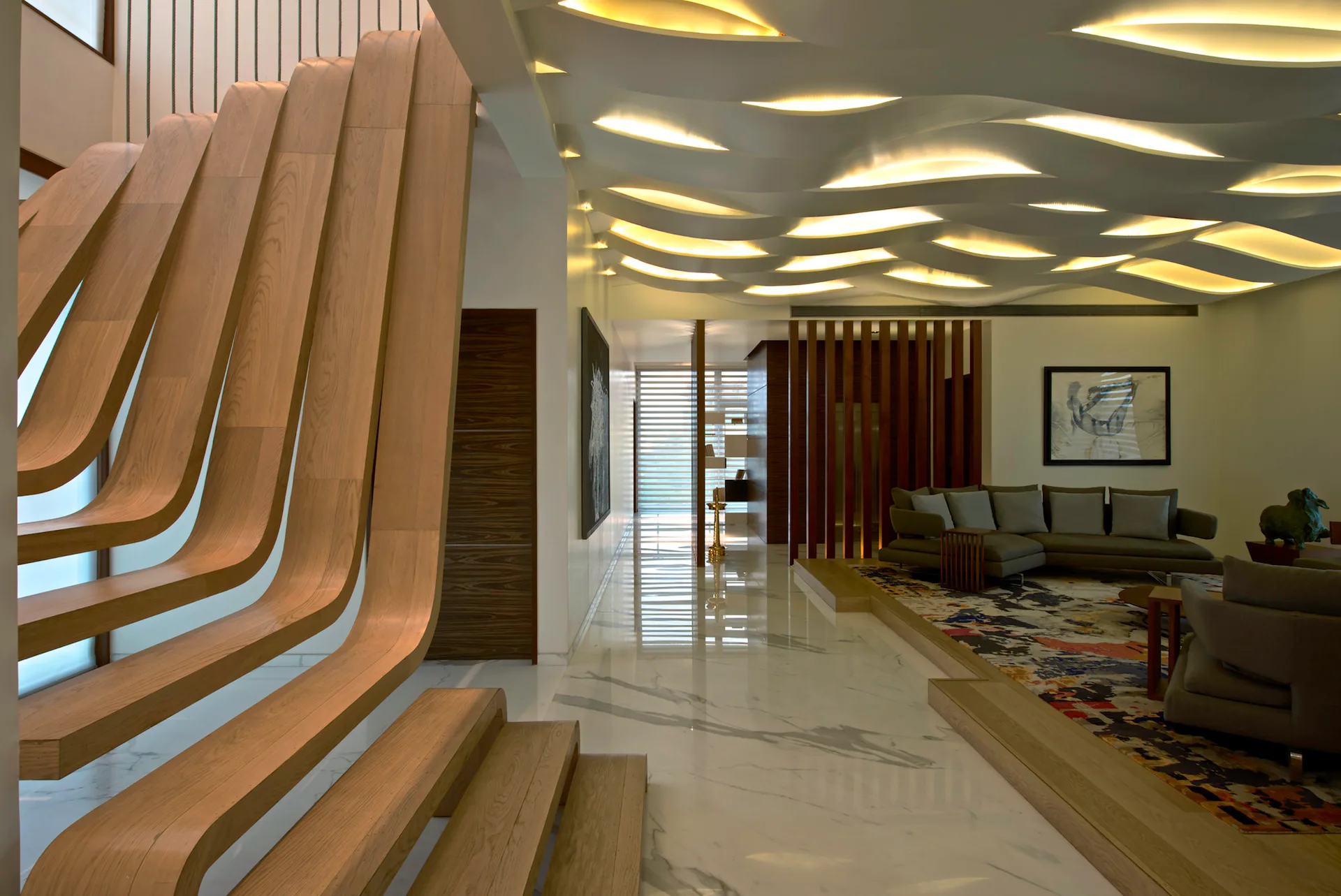 A contemporary living room featuring a sculptural curved wood staircase, a wave-patterned ceiling with recessed lights, and a sunken grey sofa area.