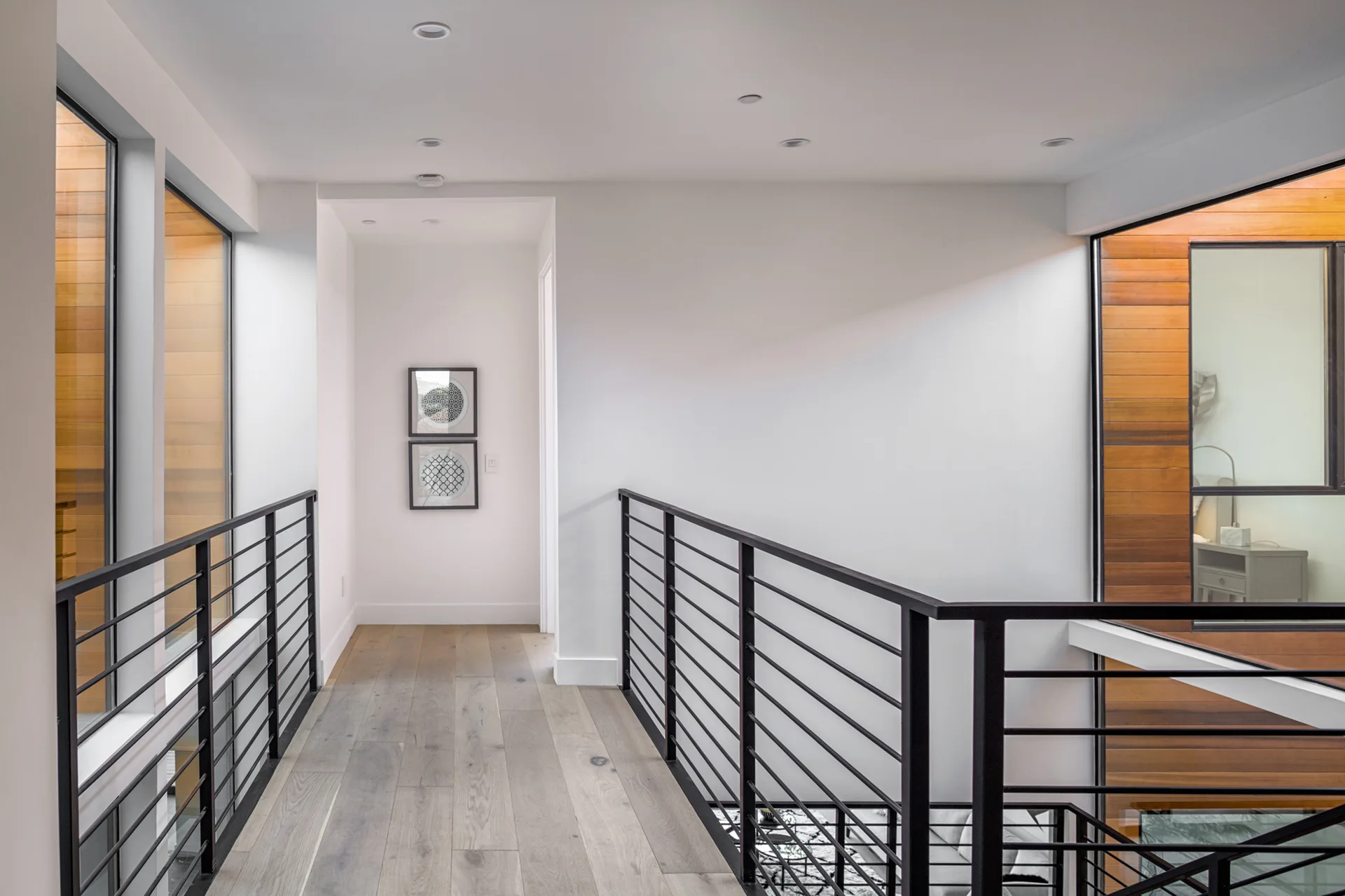 A modern second-floor hallway features light wood plank flooring, black horizontal metal railings, and white walls with stacked framed artwork.