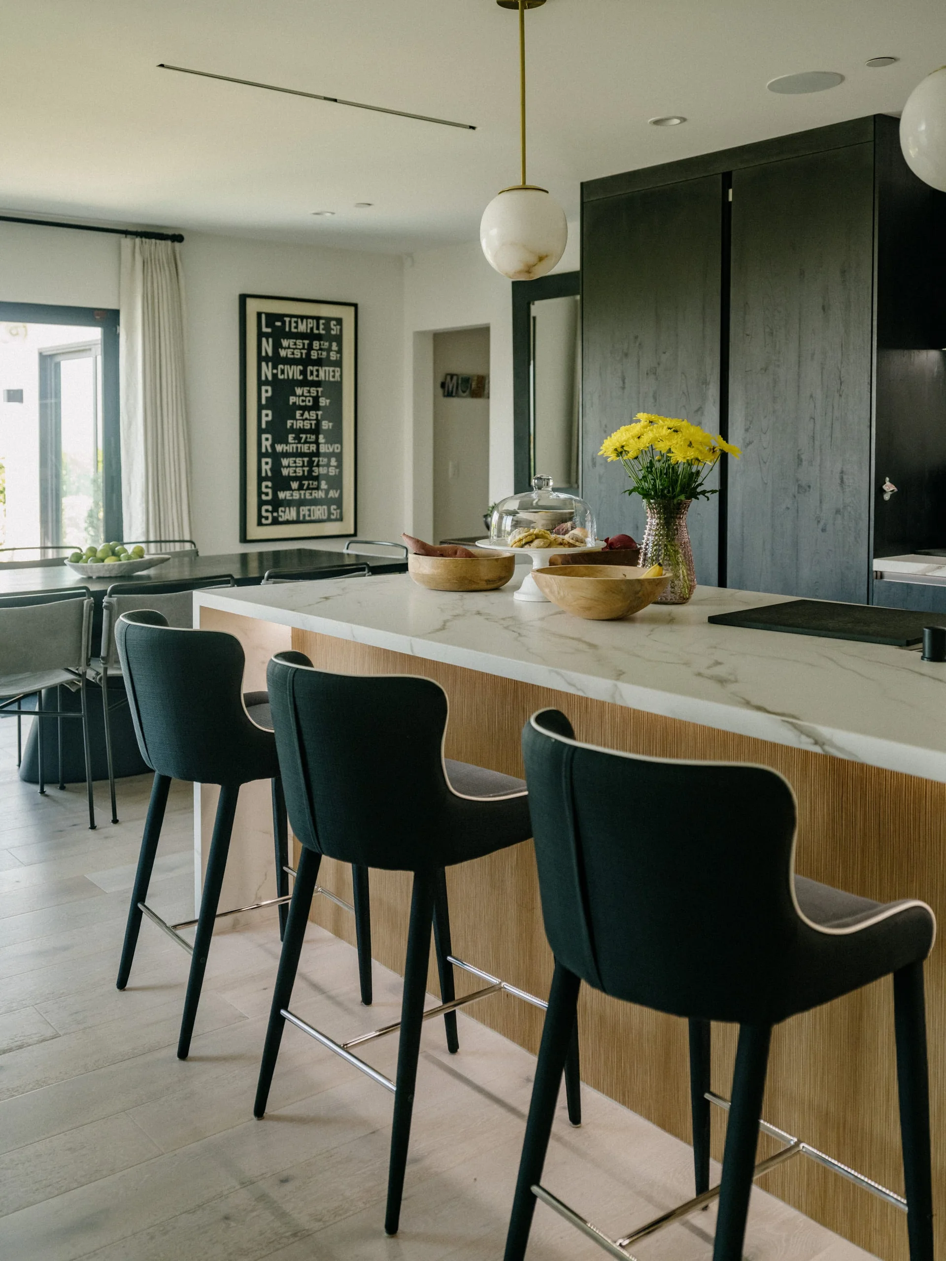 Three dark upholstered bar stools sit at a modern kitchen island featuring a veined white stone countertop and a ribbed wood base.