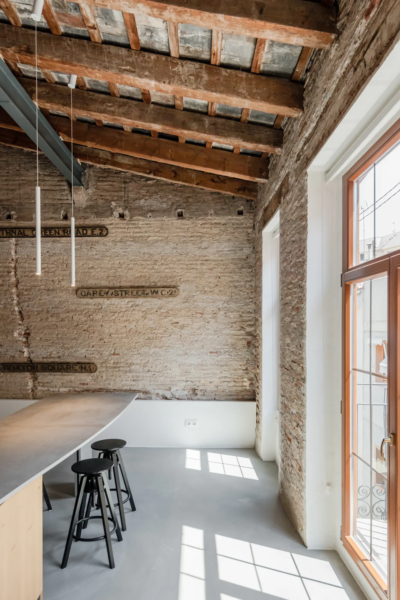 An industrial loft kitchen featuring a gray island with black stools set against a textured brick wall with vintage street signs under wooden beams.
