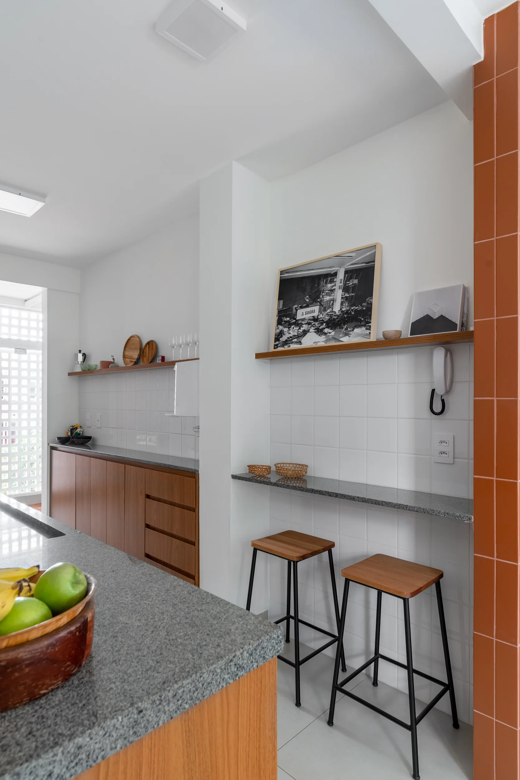 A small kitchen with wood cabinets, grey granite countertops, white square tile backsplash, wooden stools under a narrow bar, and an orange tiled wall.