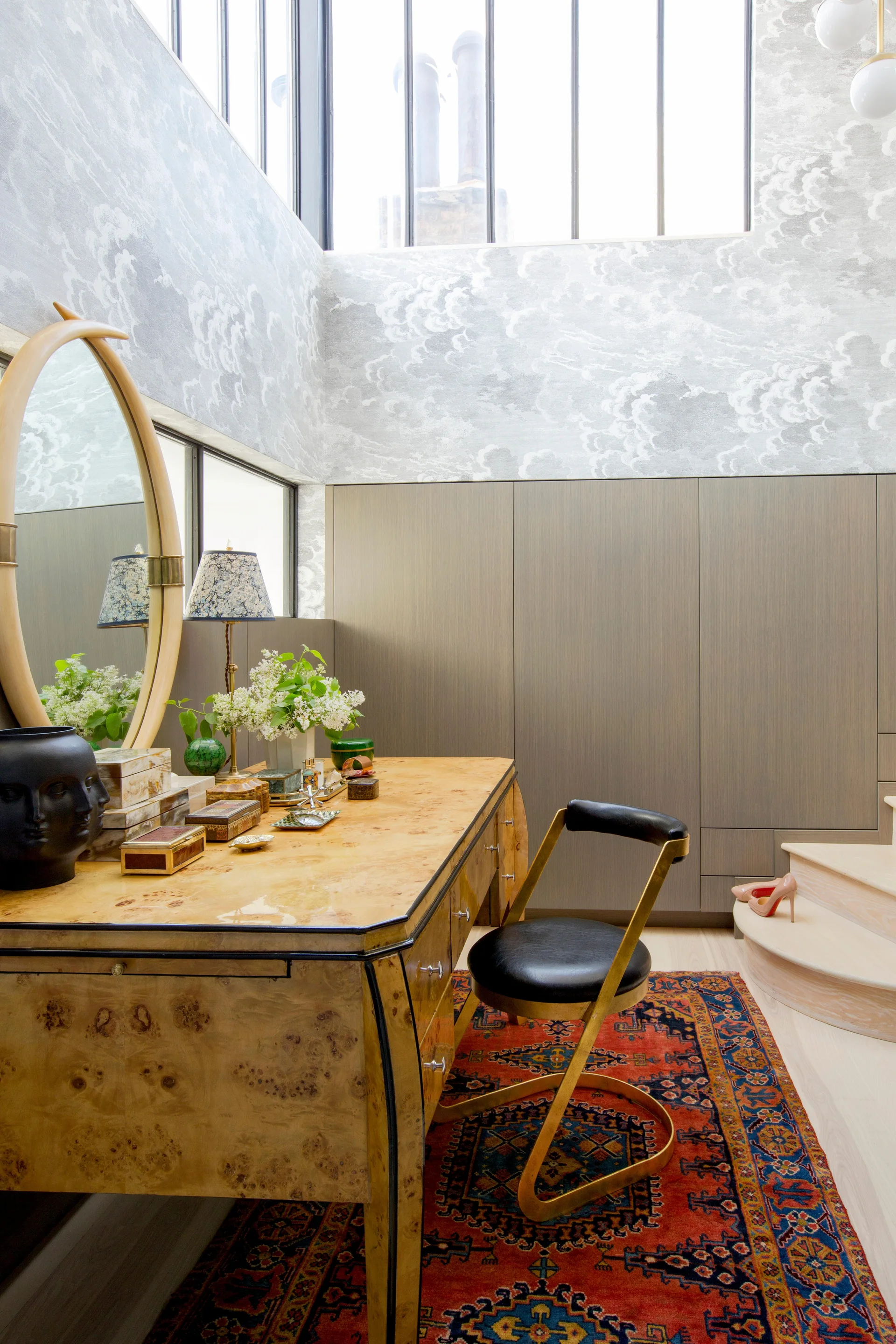 A burl wood vanity desk with an oval mirror and black chair sits on a colorful oriental rug next to built-in wardrobes and cloud-patterned wallpaper.