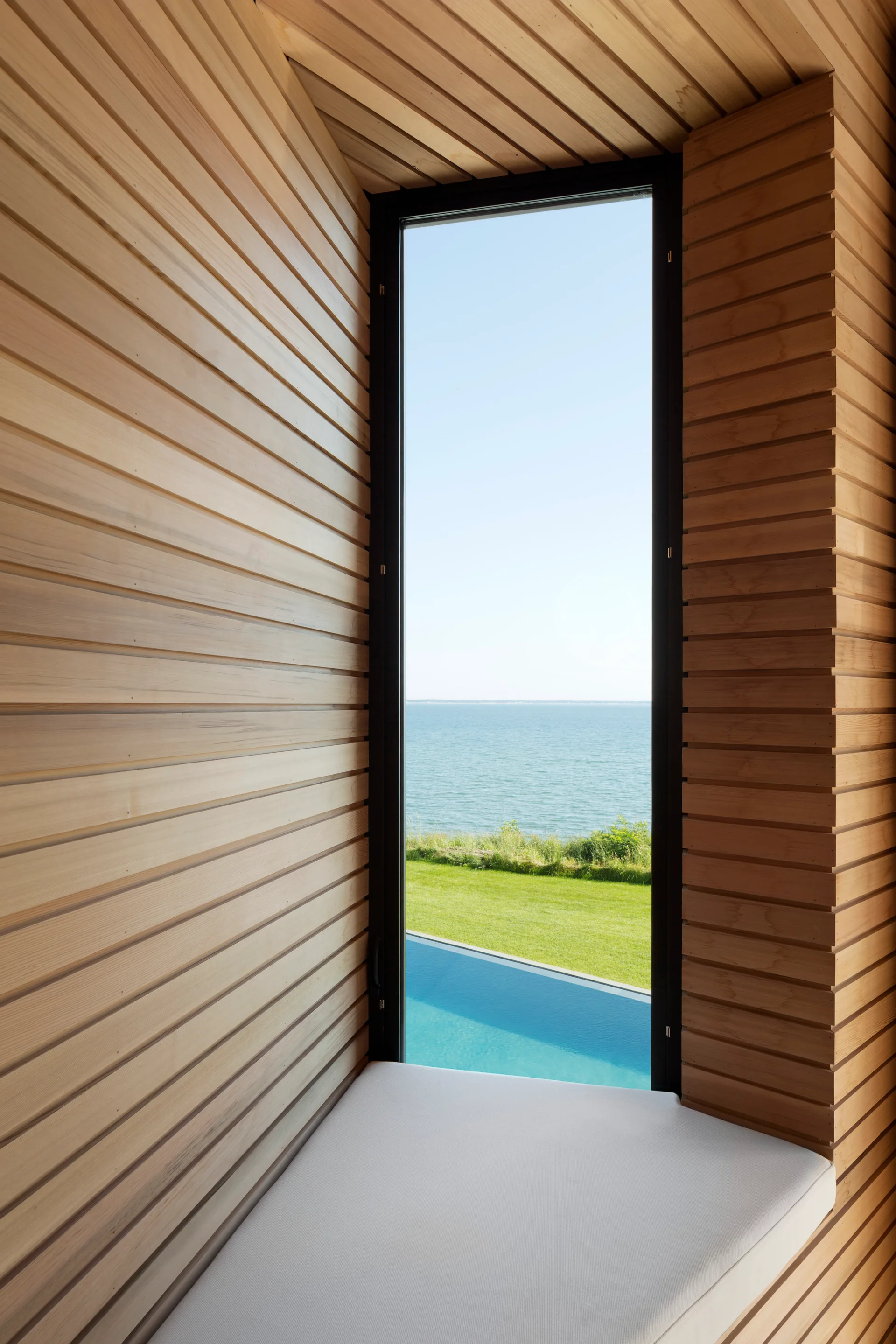 A built-in window seat with a white cushion is recessed in an alcove lined with horizontal light wood paneling, framing an ocean and pool view.
