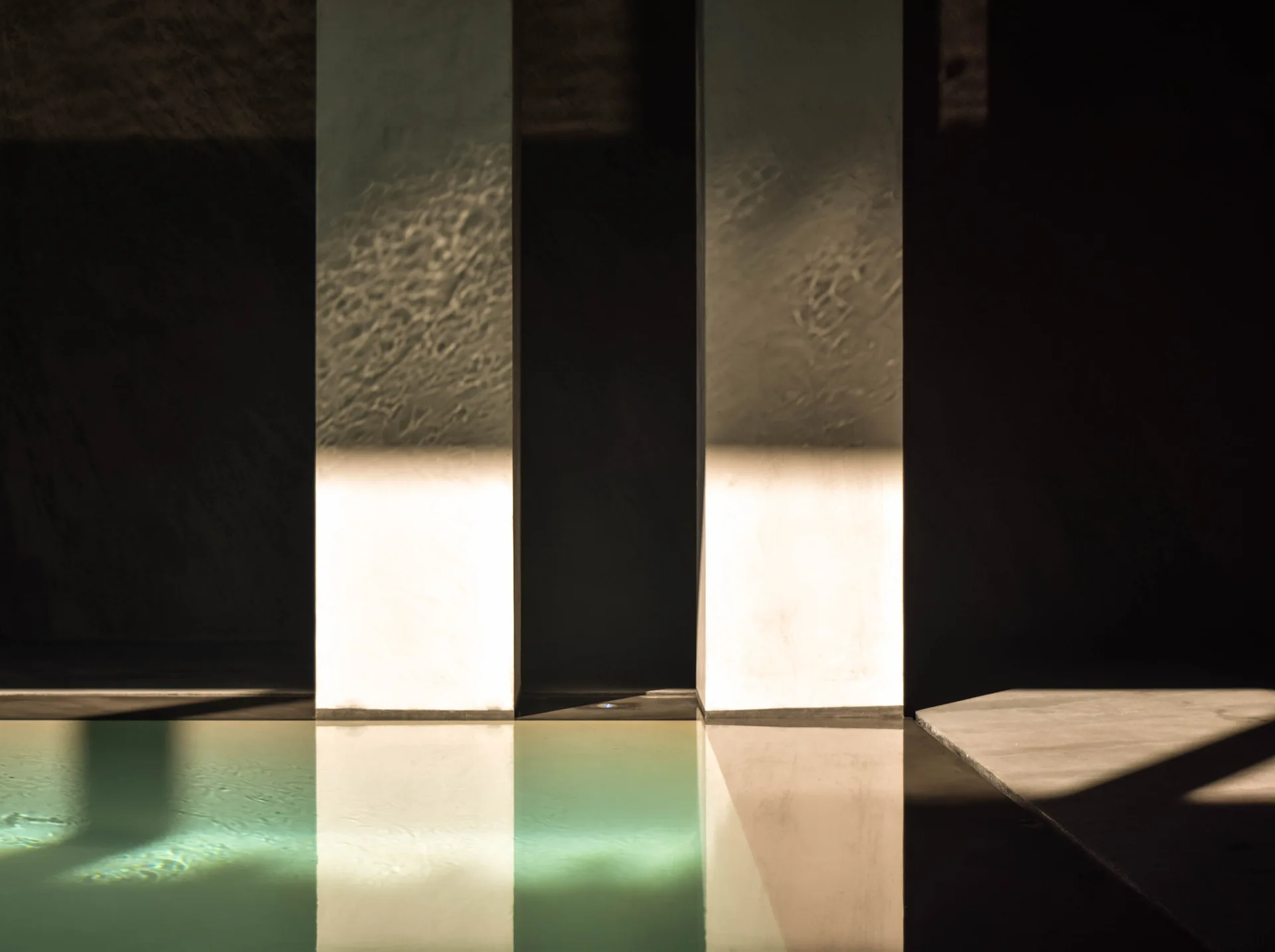 Two vertical concrete pillars stand beside a glowing turquoise swimming pool, casting sharp shadows and reflecting water ripples on their surfaces.