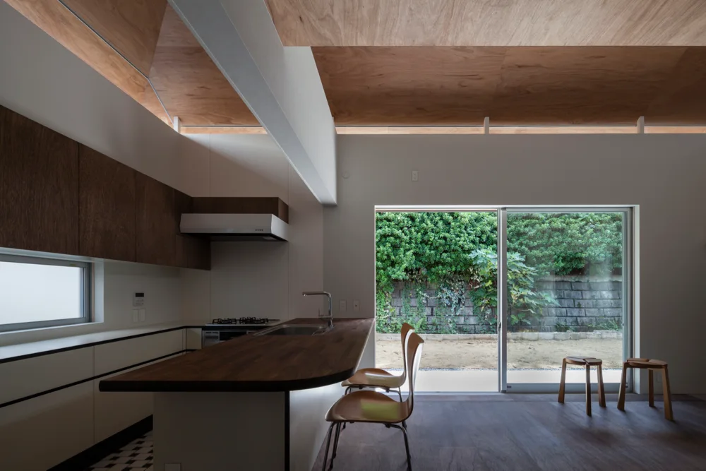 A minimalist kitchen features a dark wood counter, white lower cabinets, a sloped wood ceiling, and sliding glass doors leading to a lush garden.