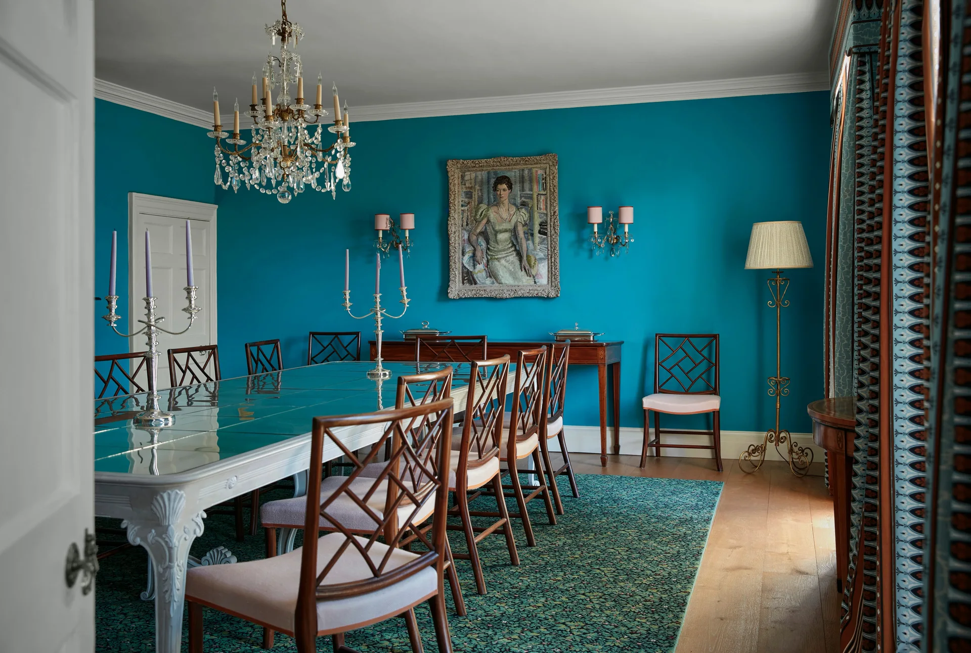 A traditional dining room featuring a glass-topped table, wooden lattice-back chairs, patterned rug, and crystal chandelier against bright teal walls.