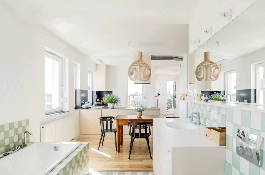 A bright open-plan space containing a white kitchen and dining area next to a bathtub and vanity detailed with green checkerboard tiles.