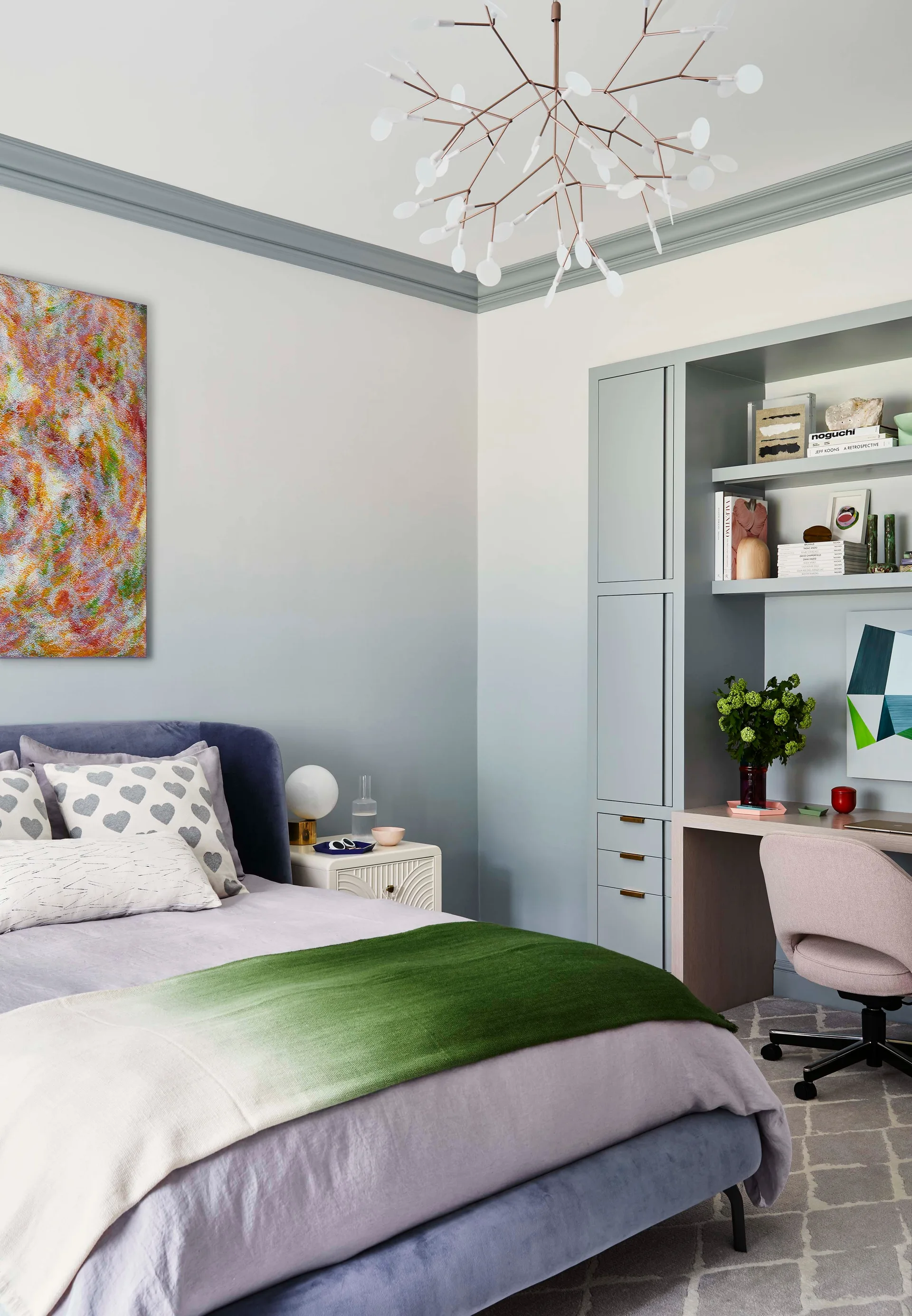 A contemporary bedroom features an upholstered blue bed, a built-in grey desk with shelving, gradient painted walls, and a branching chandelier.