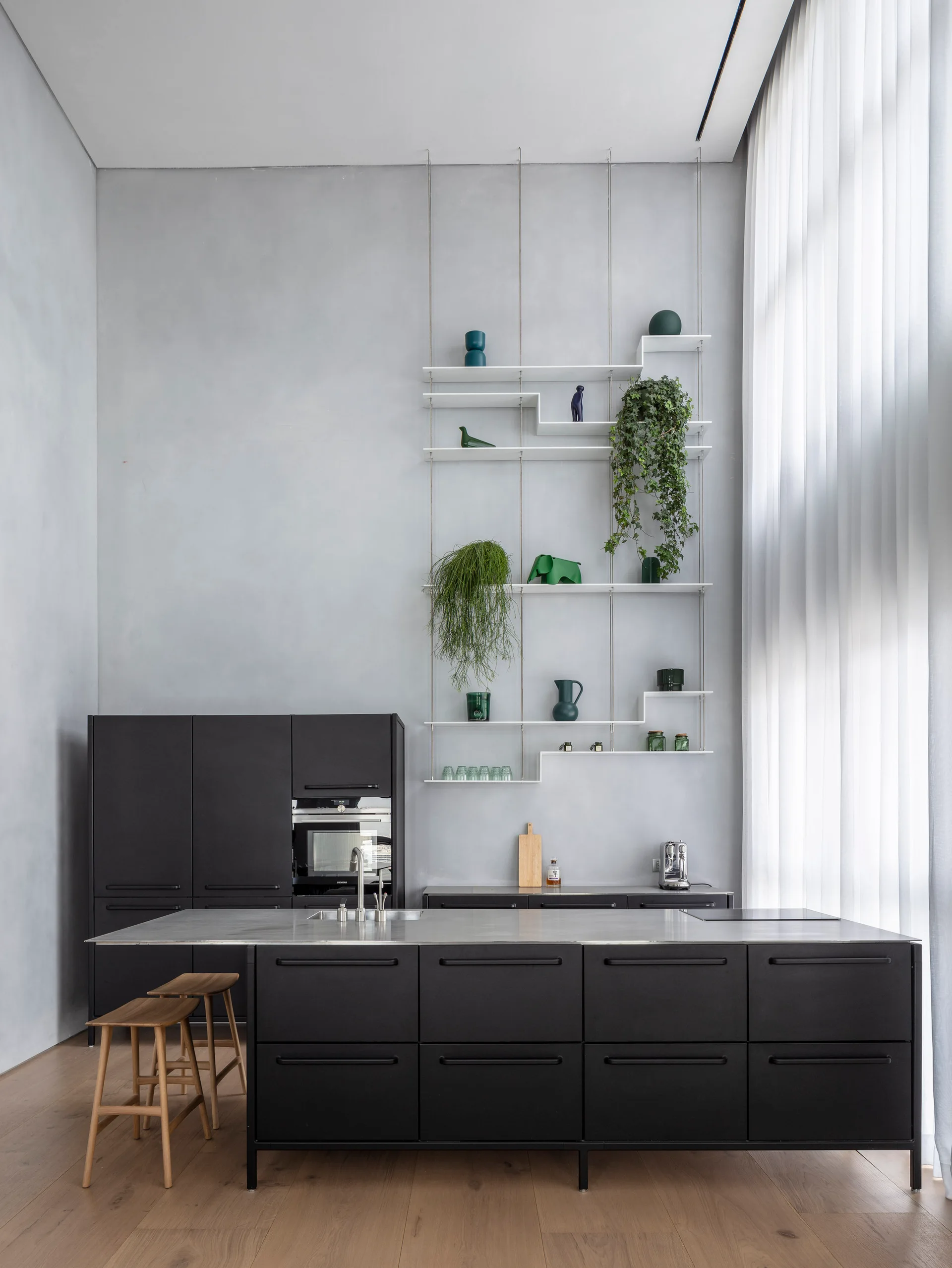 A modern kitchen featuring a matte black island, matching cabinetry, wooden stools, and tall, thin wire shelving against a light grey wall.