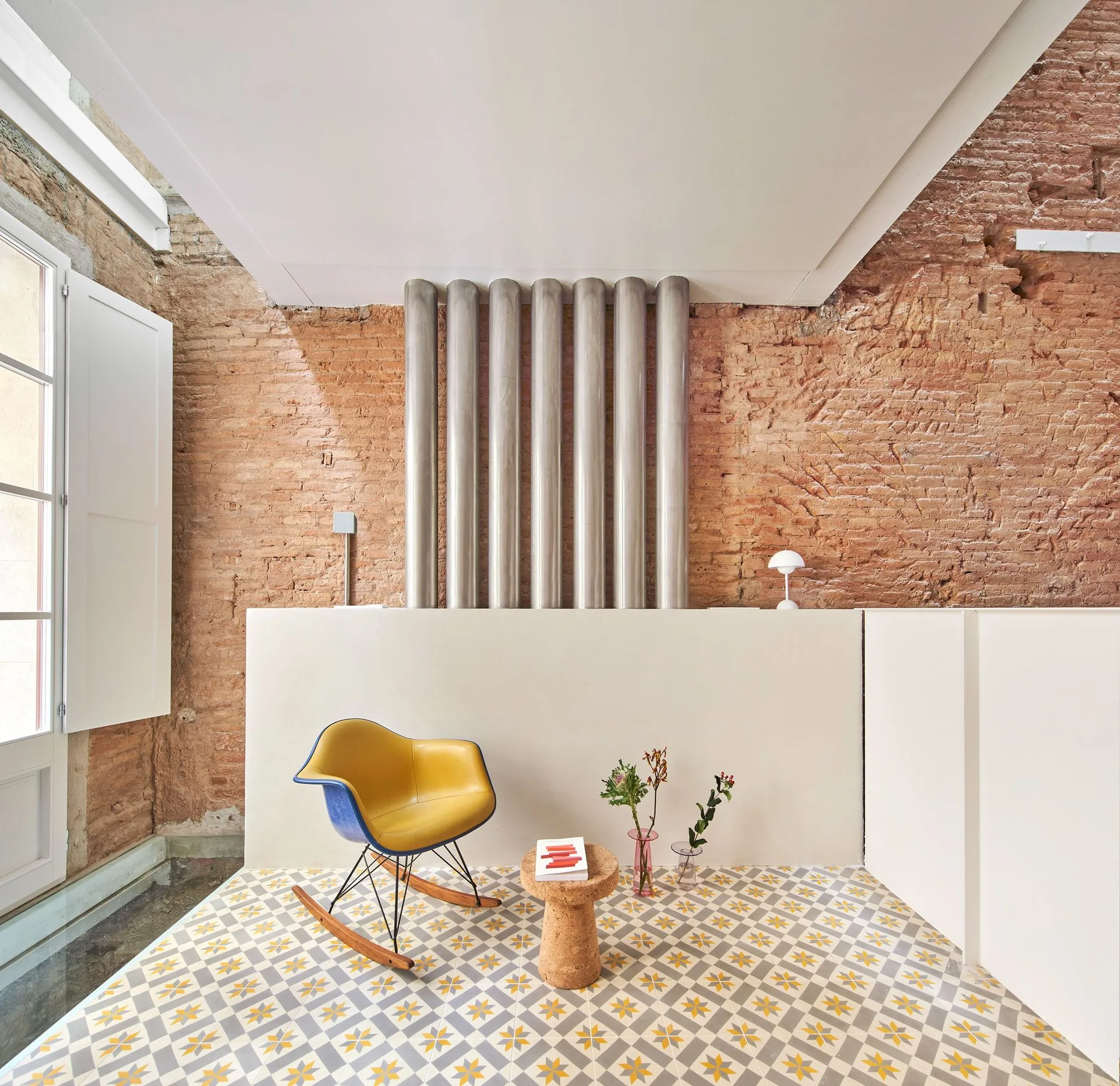 A yellow mid-century rocking chair and a cork side table sit on a yellow and grey patterned tile floor in front of an exposed brick wall.