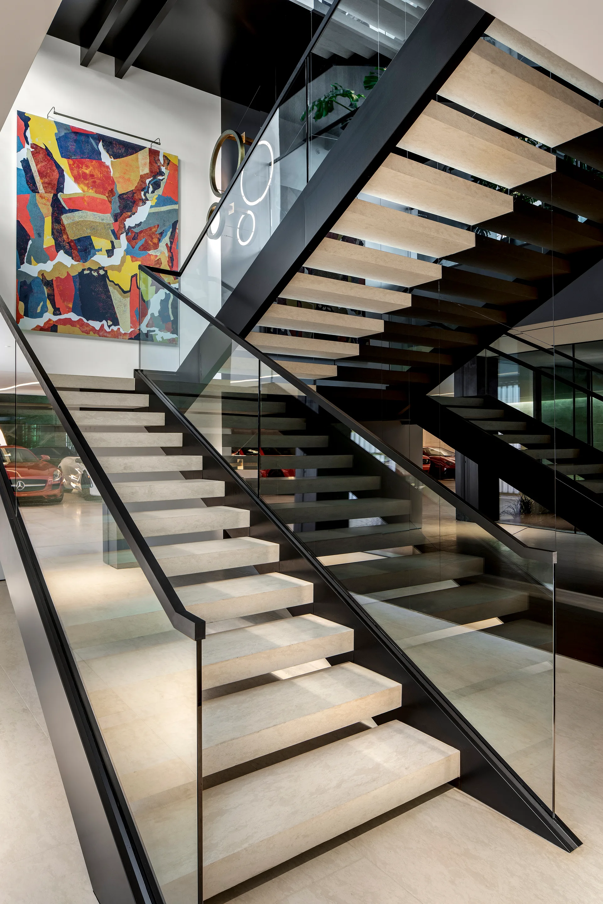 A modern floating staircase with beige stone steps, black metal trim, glass railings, and a large abstract painting on the wall.