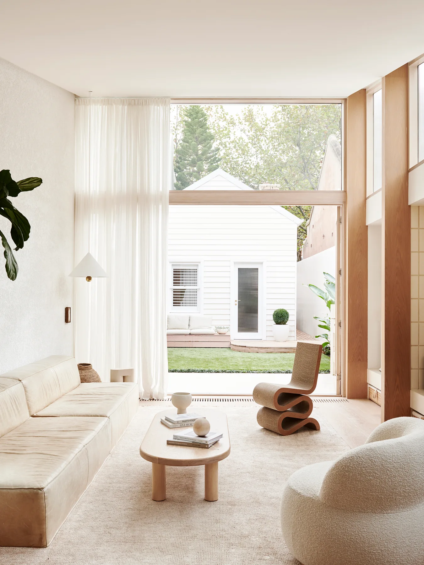 A bright living room features a cream leather sofa, an oval wooden coffee table, a sculptural wavy chair, and a large glass door opening to a garden.