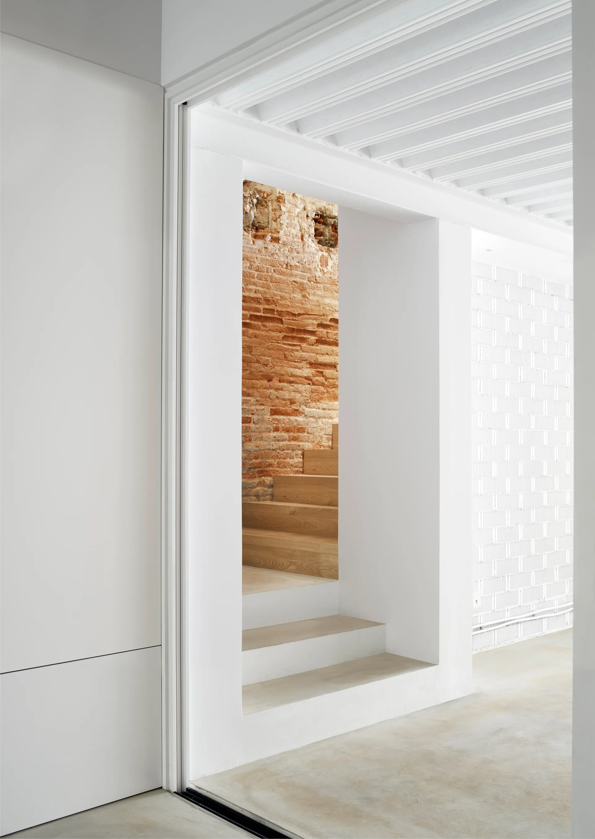 A modern wooden staircase is visible through a thick white plaster doorway against a rustic exposed terracotta brick wall.