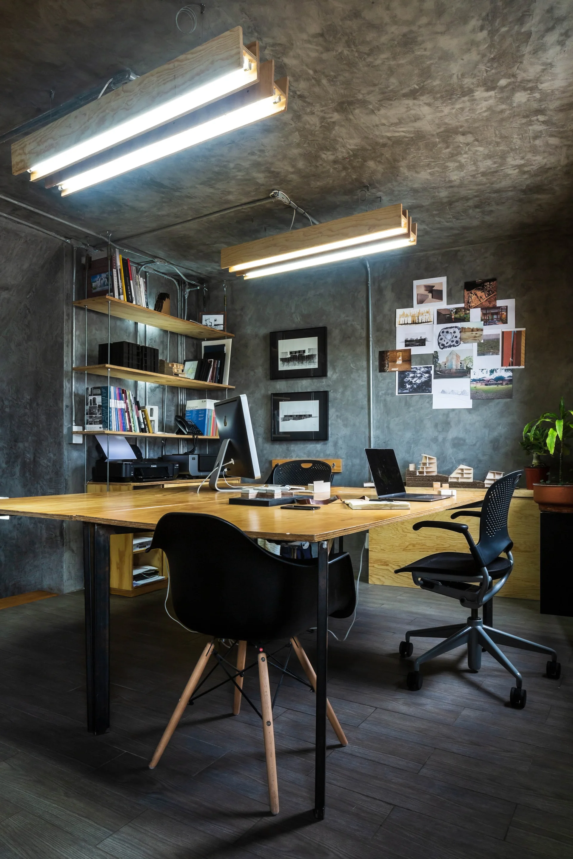 An industrial home office features a large wooden desk, black office chairs, textured concrete walls, and wooden suspended fluorescent light fixtures.