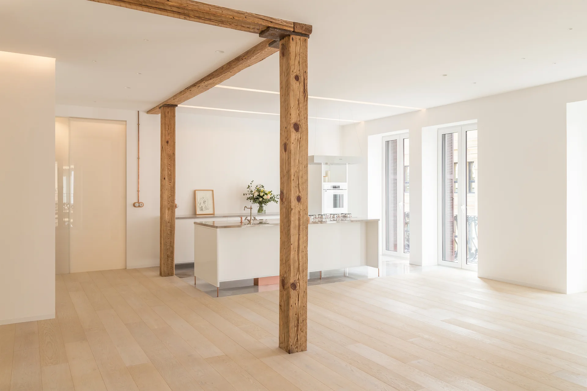A minimalist white kitchen features a central island, light wood flooring, and prominent rustic timber columns supporting a ceiling beam.