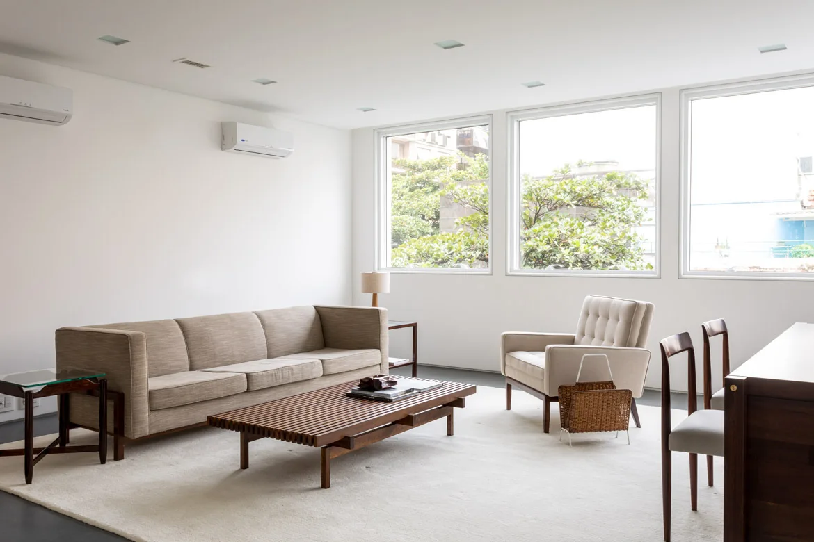 A bright living room features a beige sofa, a slatted wood coffee table, and an armchair arranged on a cream rug.