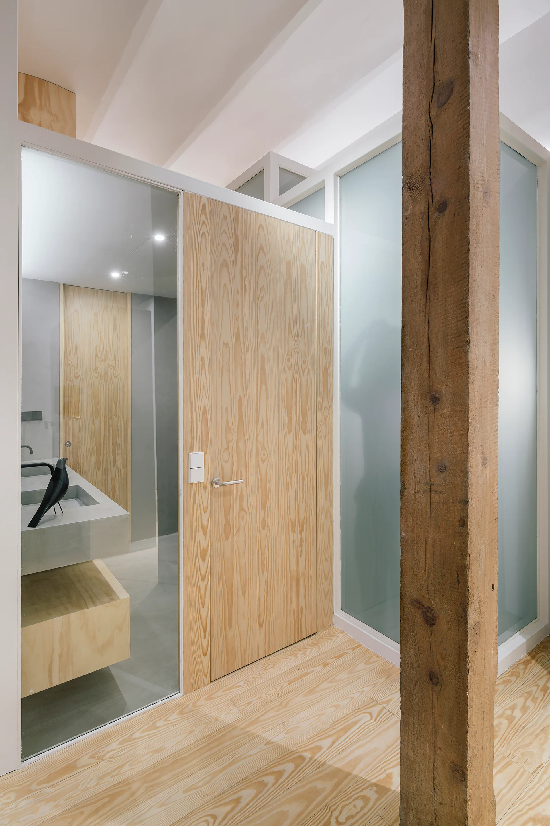 A modern bathroom entry features a pale wood door, frosted glass partitions, a concrete vanity, and a rustic structural timber column.