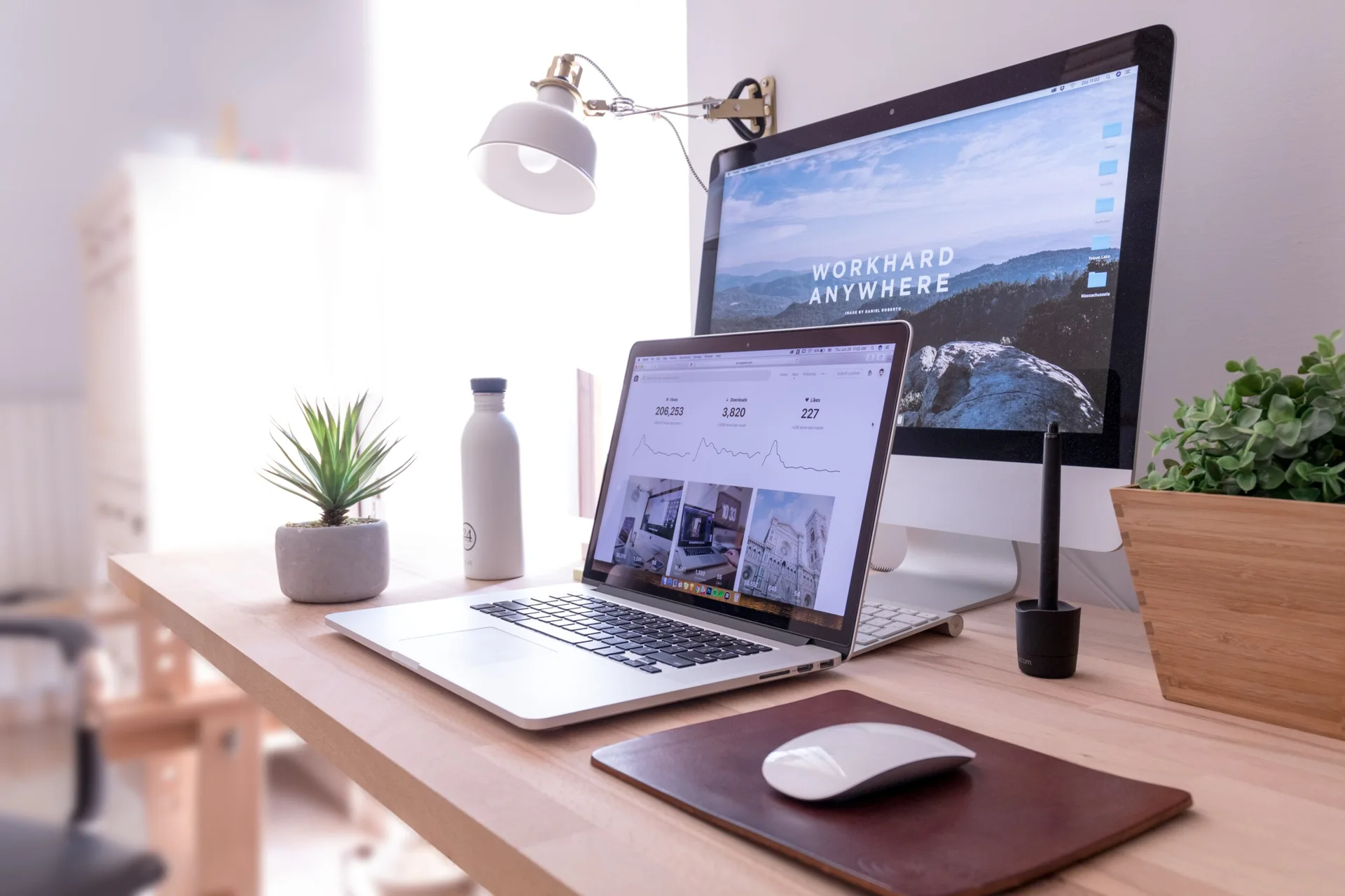 A light wood home office desk features a laptop, desktop computer, small potted plants, and a wall-mounted white reading lamp.