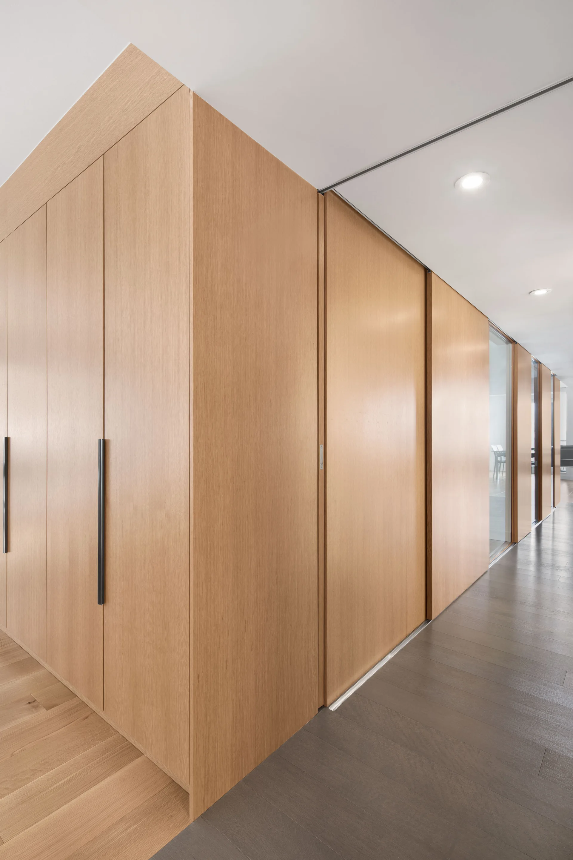 Floor-to-ceiling wooden cabinets with minimalist black handles line a clean hallway corridor featuring recessed lighting and wood floors.