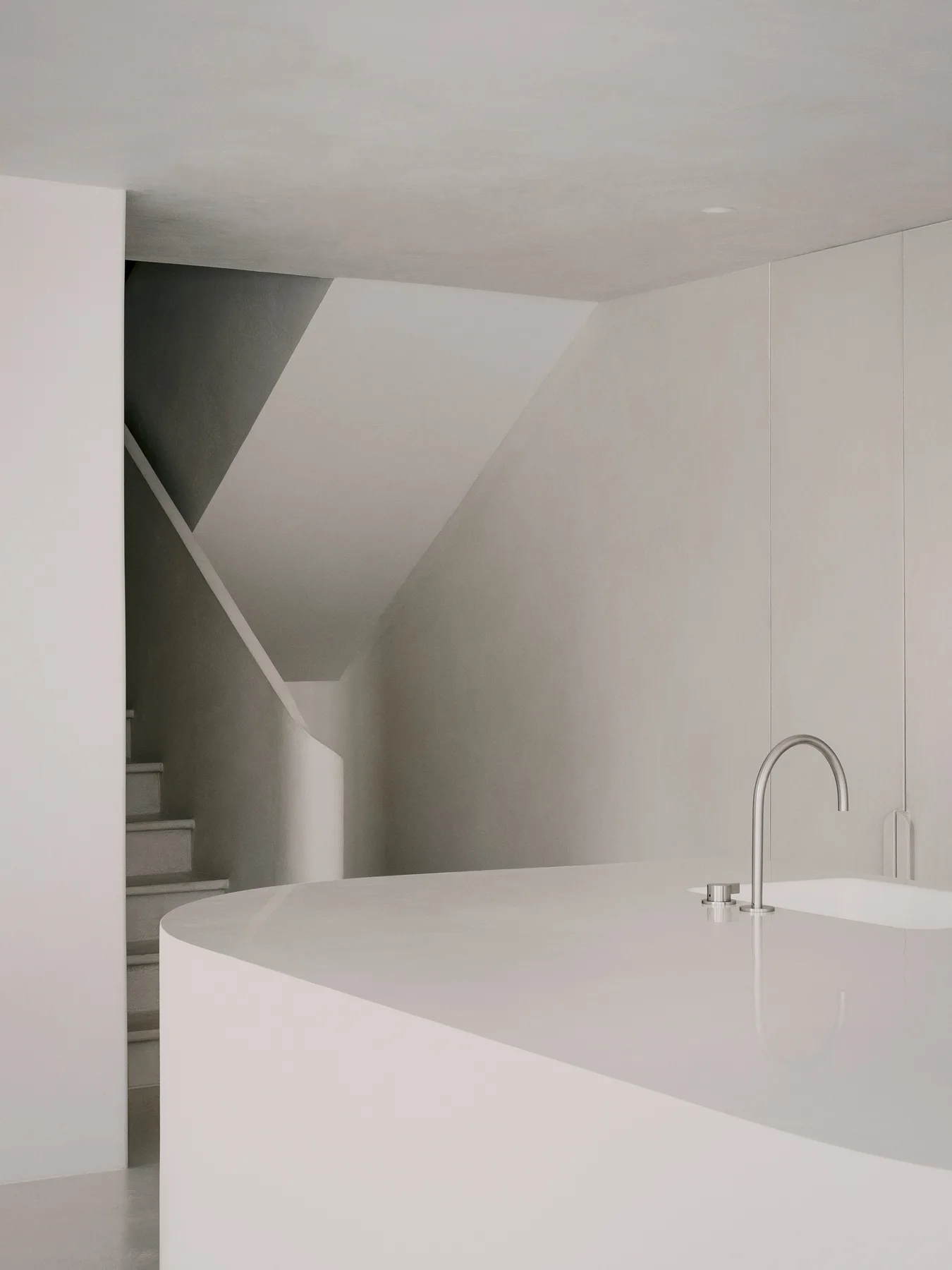 A minimalist off-white bathroom features a curved vanity with a metal faucet in front of a seamless plaster staircase.