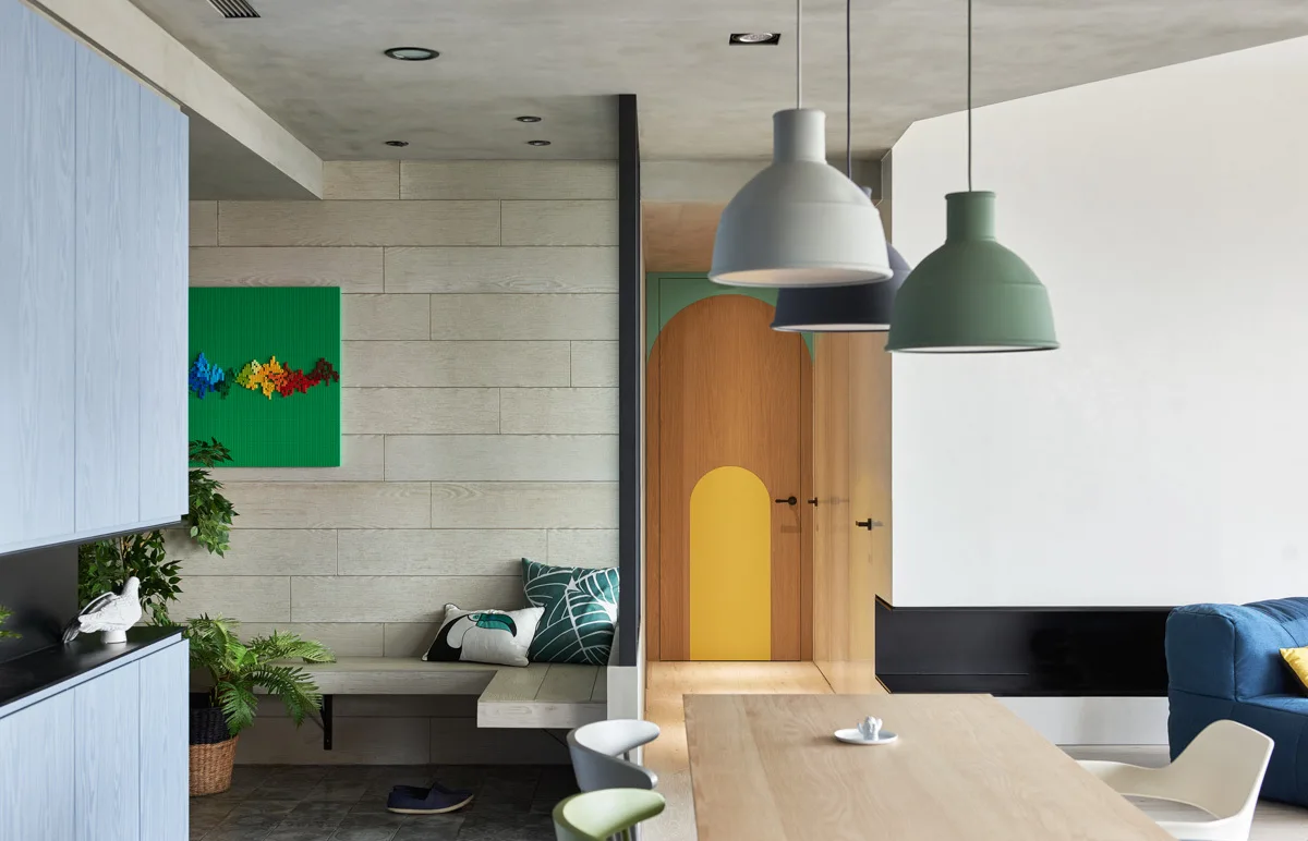 A modern dining area features a light oak table, three hanging pendant lights, and a wooden door with painted arches at the end of a hallway.