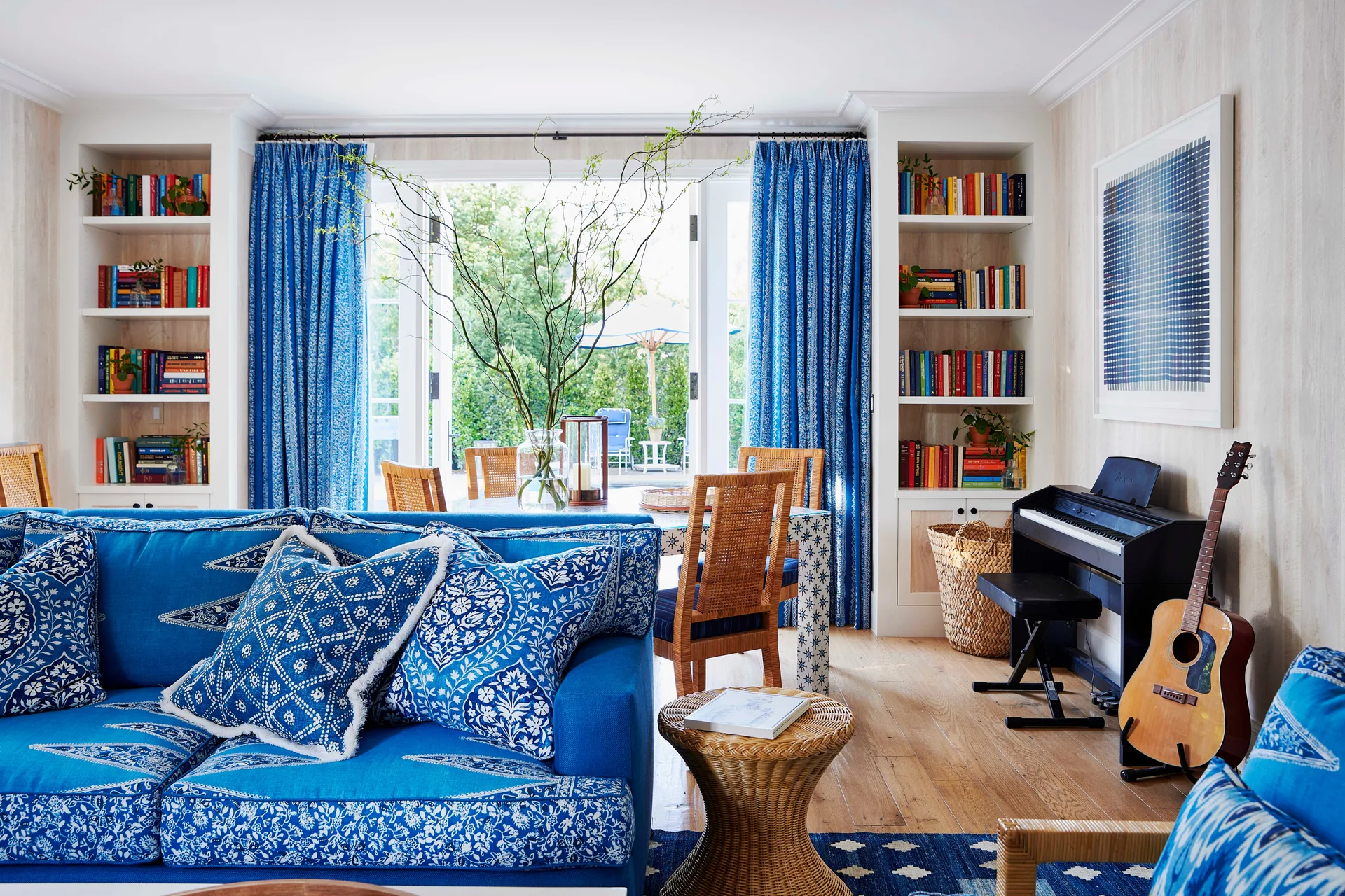 A bright living room features a blue patterned sofa, symmetrical built-in bookshelves, and open French doors leading to an outdoor patio.