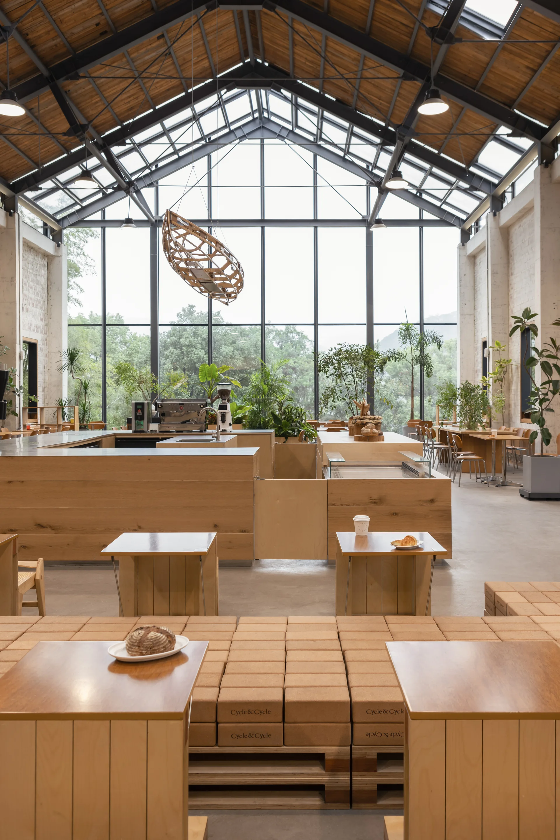 An industrial cafe interior features a high vaulted wood ceiling, modular wooden block seating, and a large glass facade showing green trees.