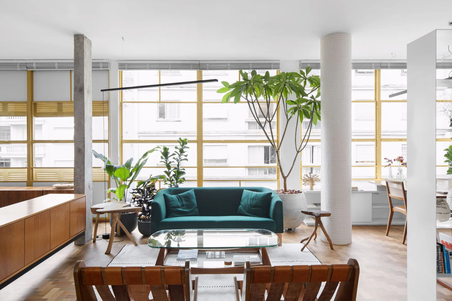 A bright modern living room features a dark green sofa, glass coffee table, and wood parquet flooring in front of large gold-framed windows.