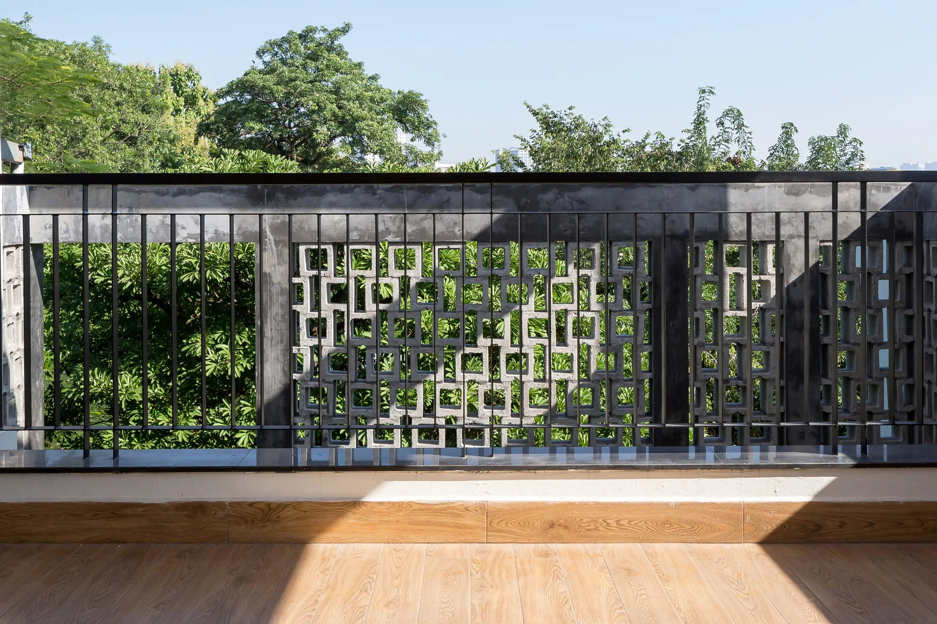 An outdoor balcony features wood-look plank flooring, a black metal railing, and decorative grey concrete breeze blocks overlooking green trees.