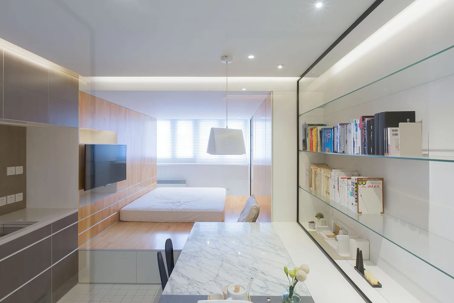 A modern studio apartment featuring a bed on a wooden platform next to a marble dining table and built-in glass bookshelves.