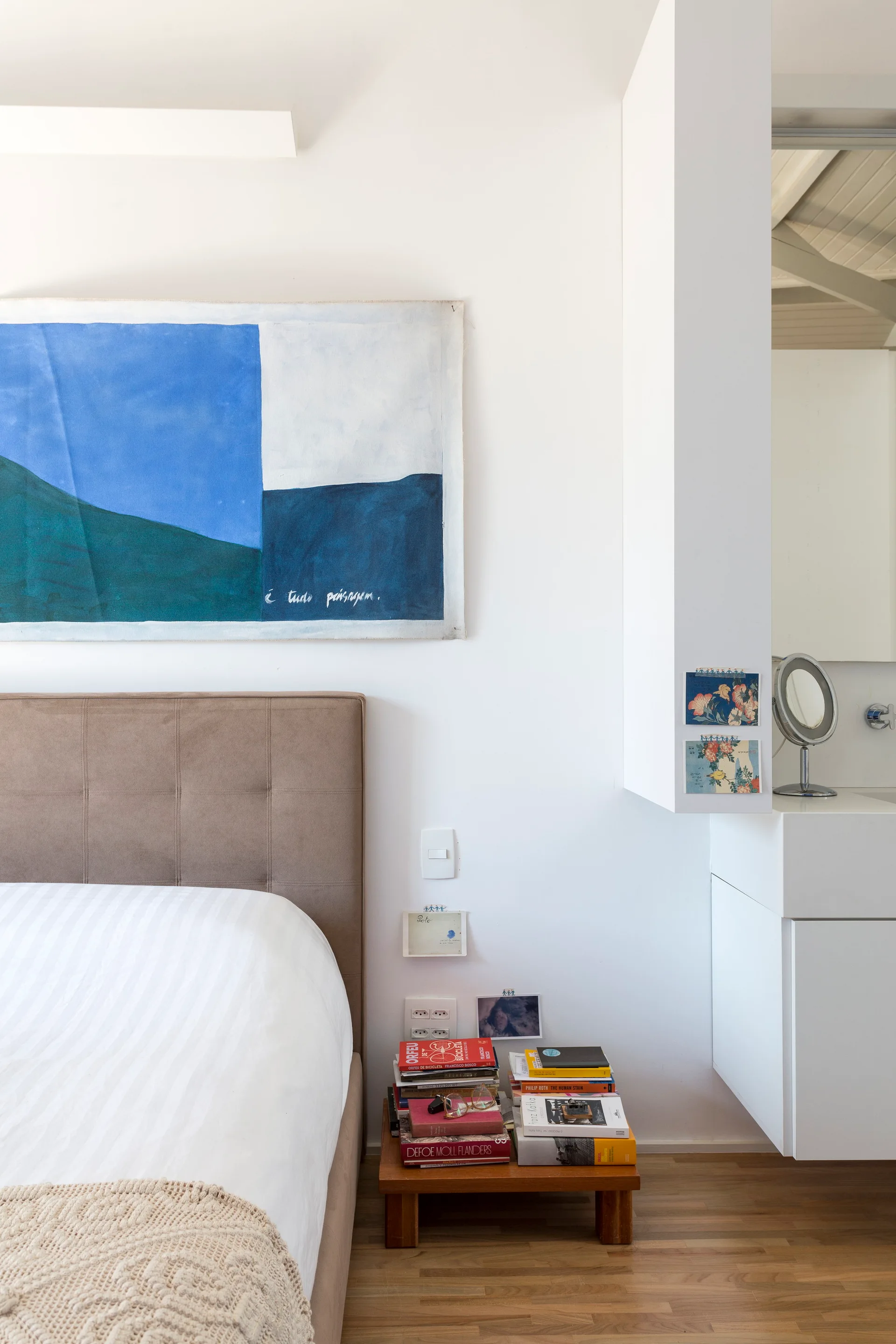 A modern bedroom features a brown tufted headboard, a low wooden nightstand stacked with books, and an integrated white vanity cabinet.