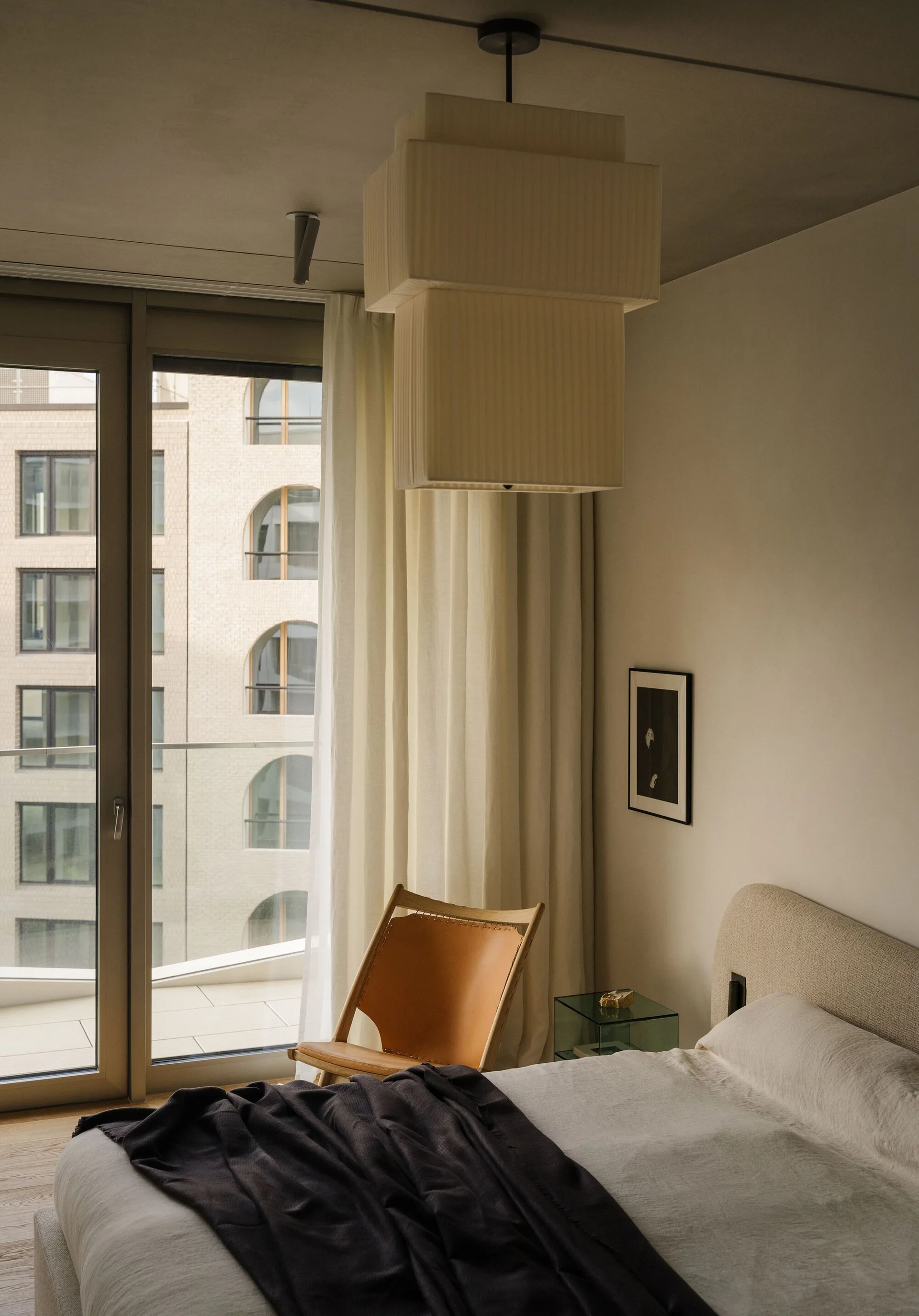 A modern bedroom features a low bed with beige linens, a tan leather sling chair by a balcony window, and a large tiered fabric pendant light.