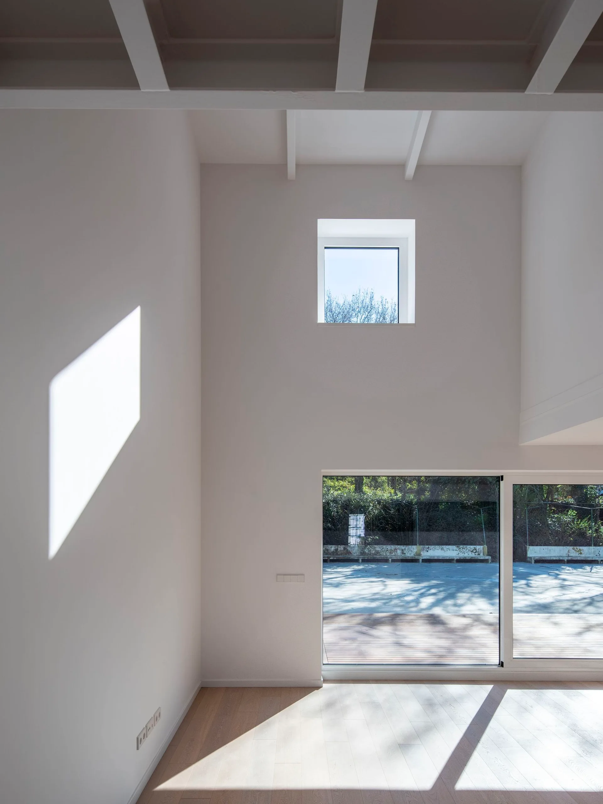 A minimalist white room features exposed ceiling beams, light wood floors, a sliding glass door, and a high square window casting a sunlight pattern.