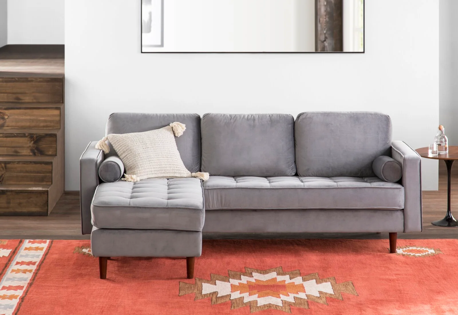 A gray velvet tufted sectional sofa with a tassel pillow sits on a rust-orange patterned area rug in front of a white wall.