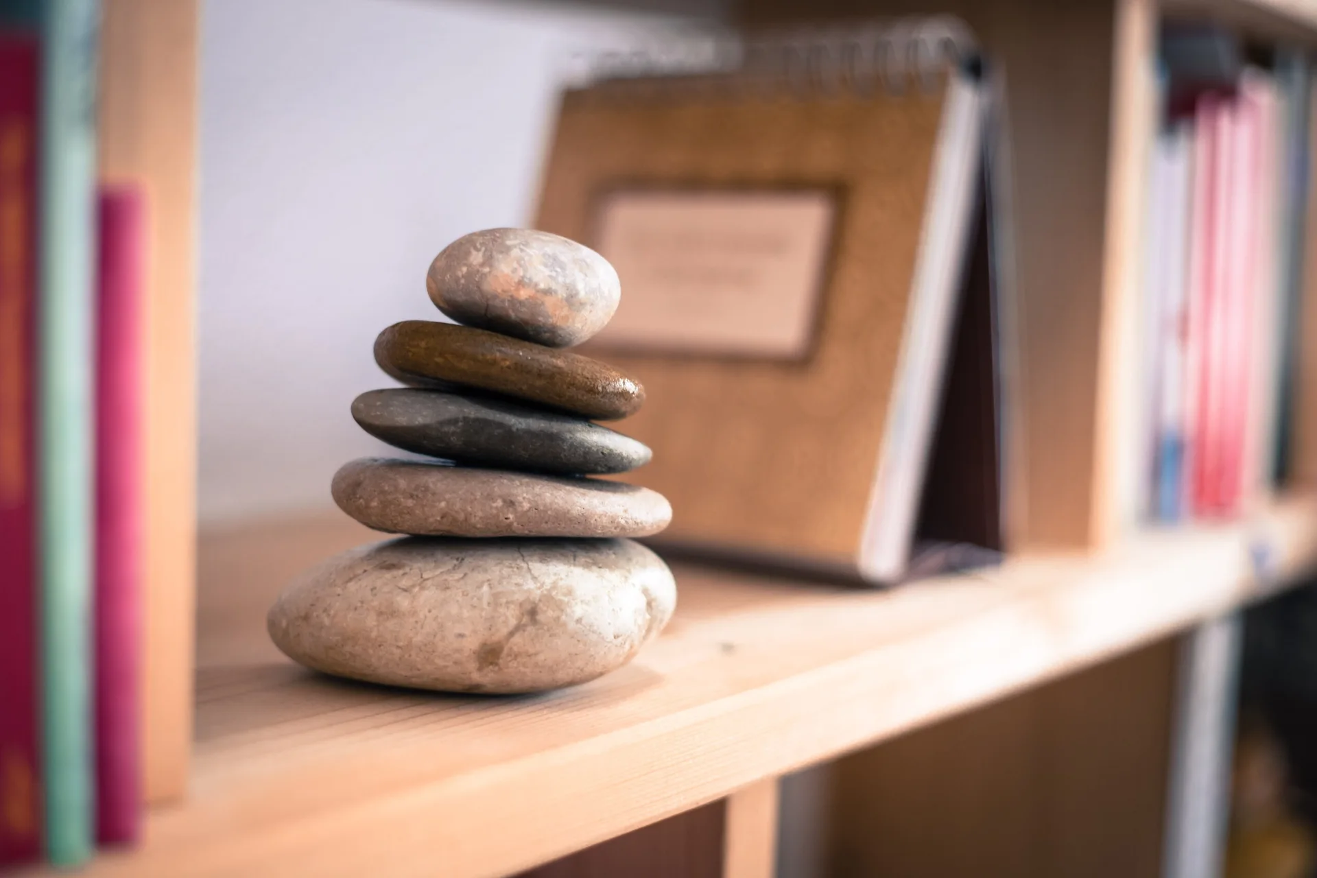 A small stack of five smooth, rounded river stones in grey and tan tones sits on a light wood bookshelf with blurred books in the background.