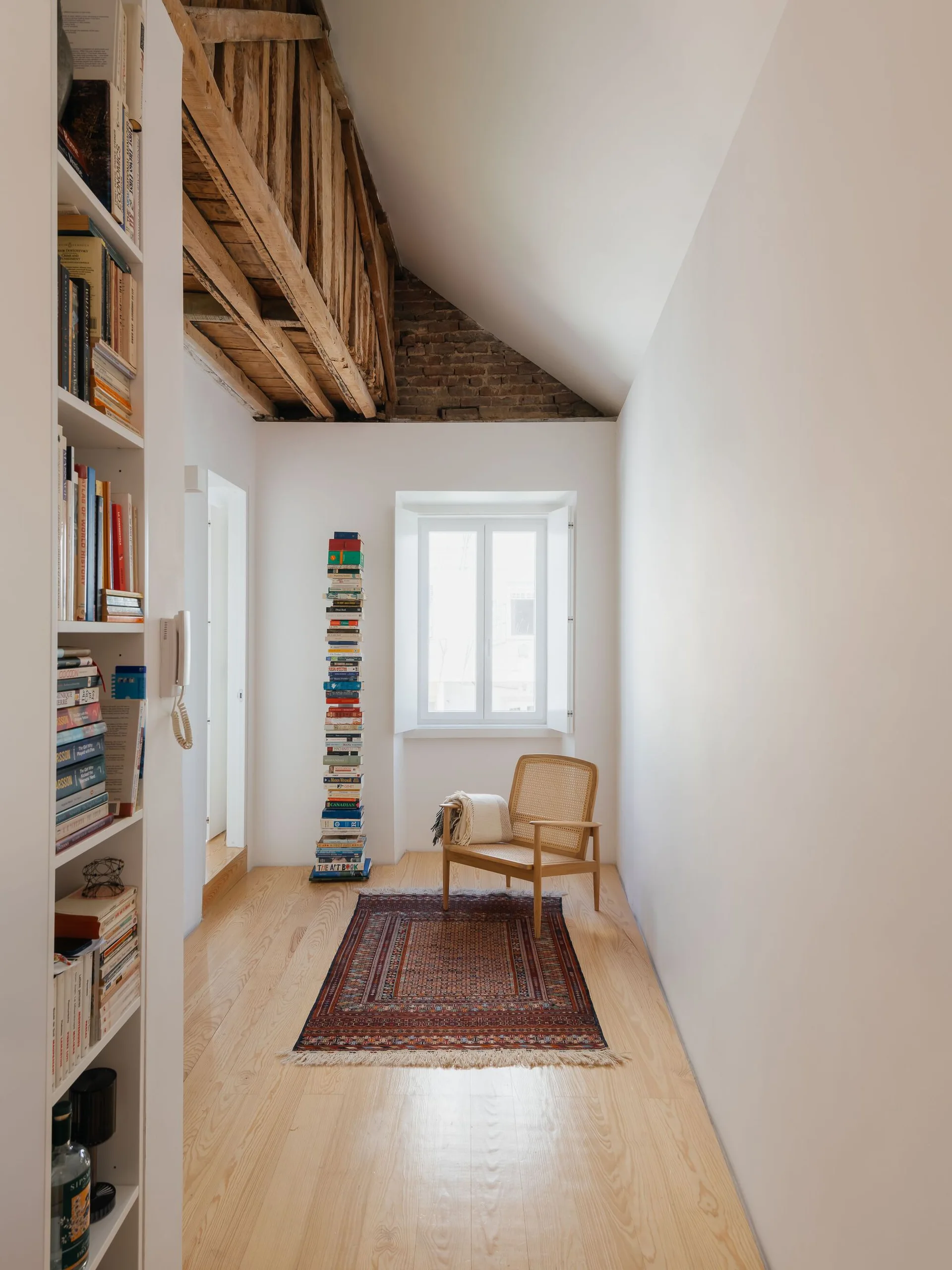 A narrow room with light wood floors features a cane armchair on a patterned rug near a window, with a tall book tower and exposed ceiling beams.