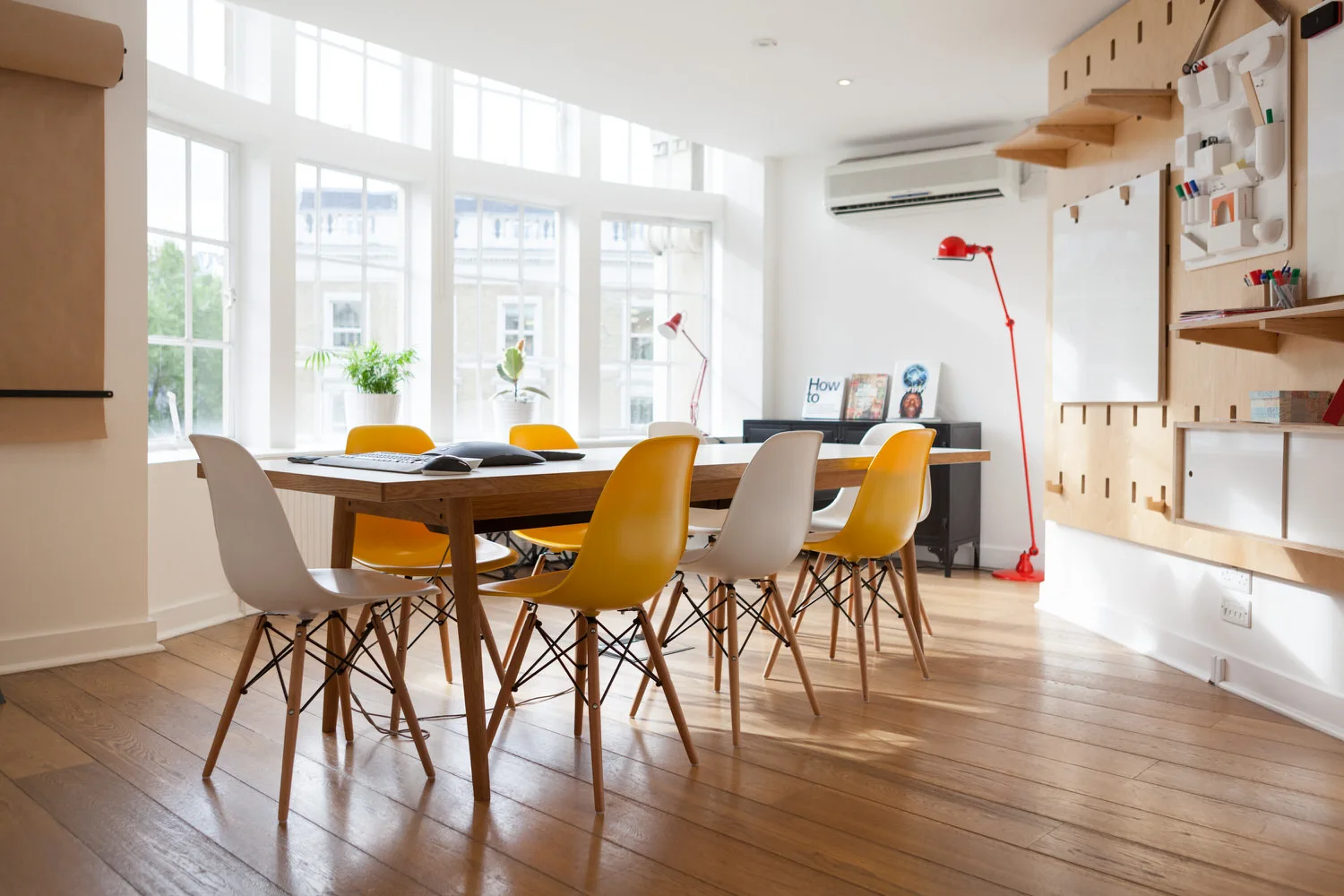 A bright workspace features a wooden conference table with yellow and white Eames-style chairs next to a large plywood wall pegboard.