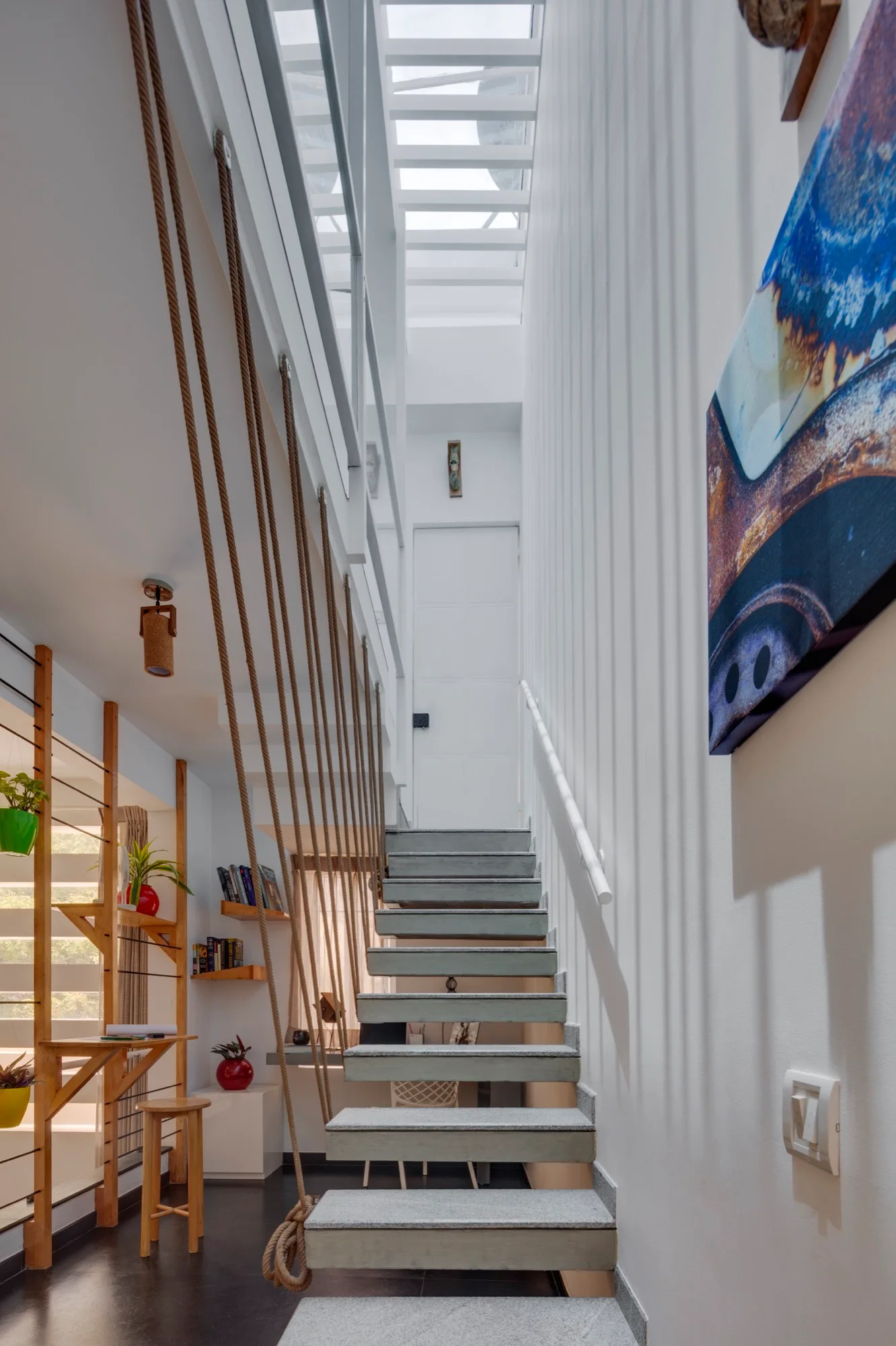 A contemporary staircase with grey stone treads and a vertical rope railing sits beneath a bright skylight in a white hallway.