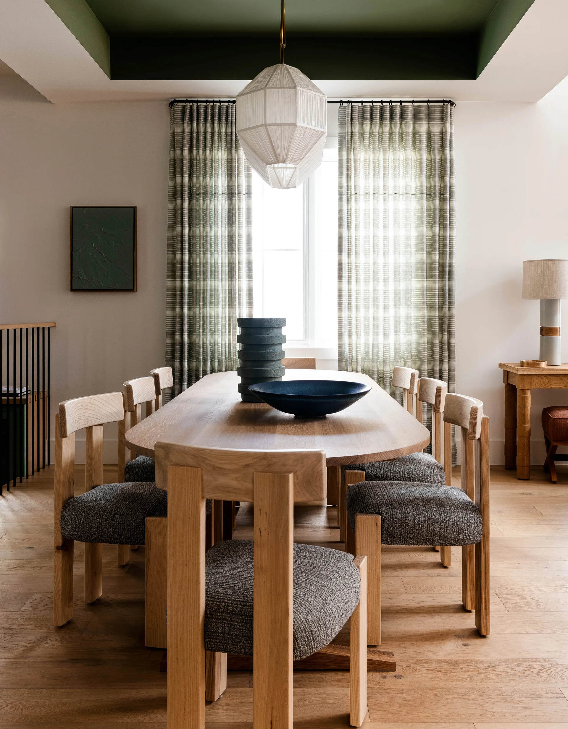 An oval wooden dining table with upholstered chairs sits beneath a geometric pendant lamp and an olive green painted tray ceiling.