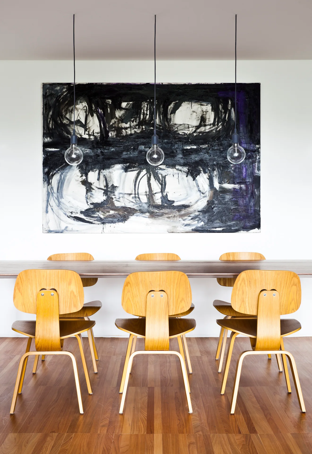 A modern dining room features six molded plywood chairs around a wooden table beneath three pendant lights and a large black and white abstract painting.