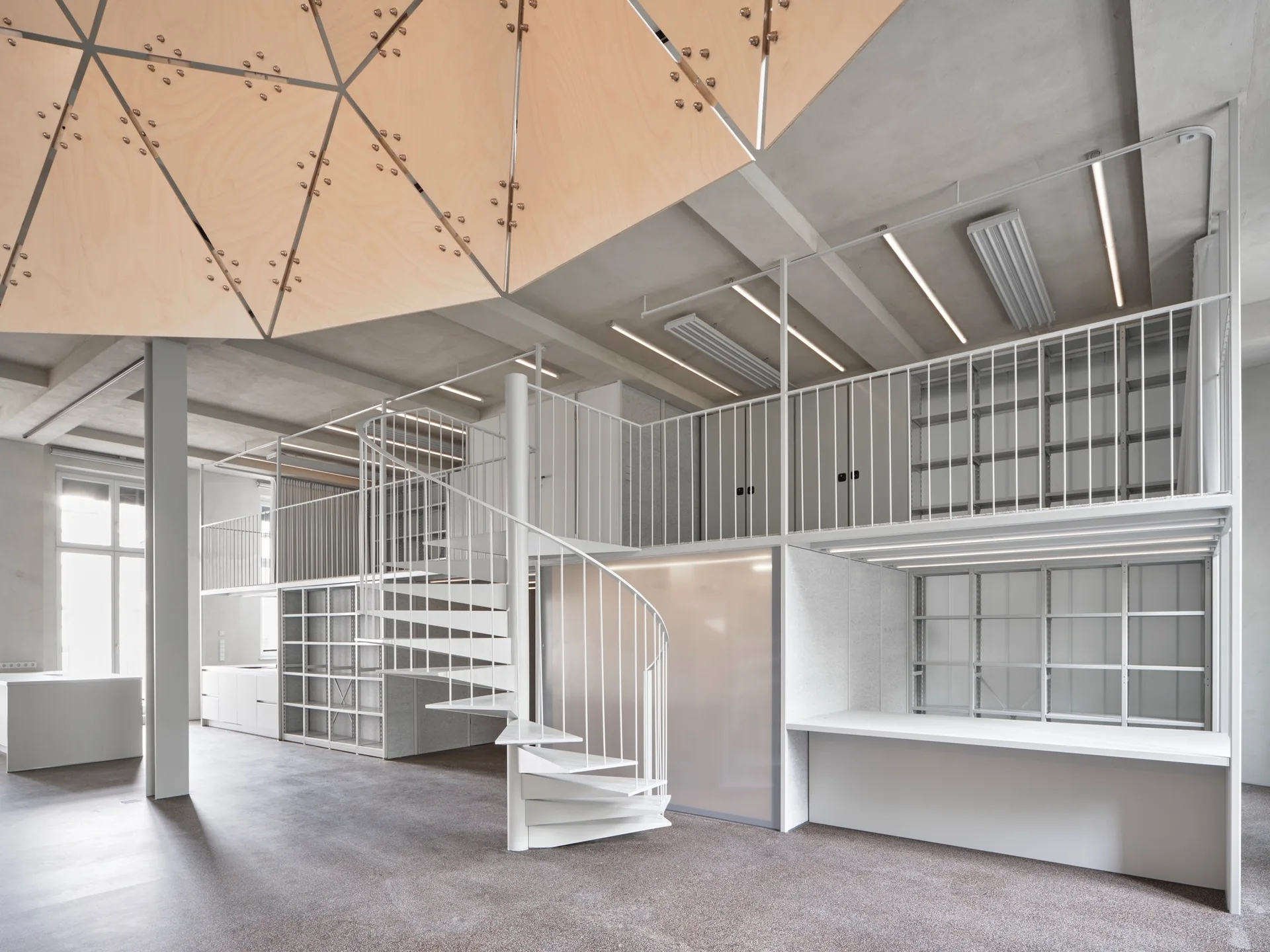 A white metal spiral staircase leads to a mezzanine level with thin railings inside an industrial room featuring a geometric wooden ceiling installation.