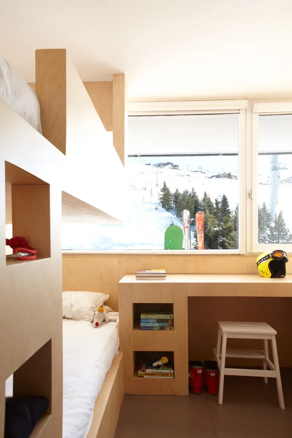 A minimalist built-in plywood bunk bed and desk unit in a kids room with a large window overlooking a snowy ski slope.