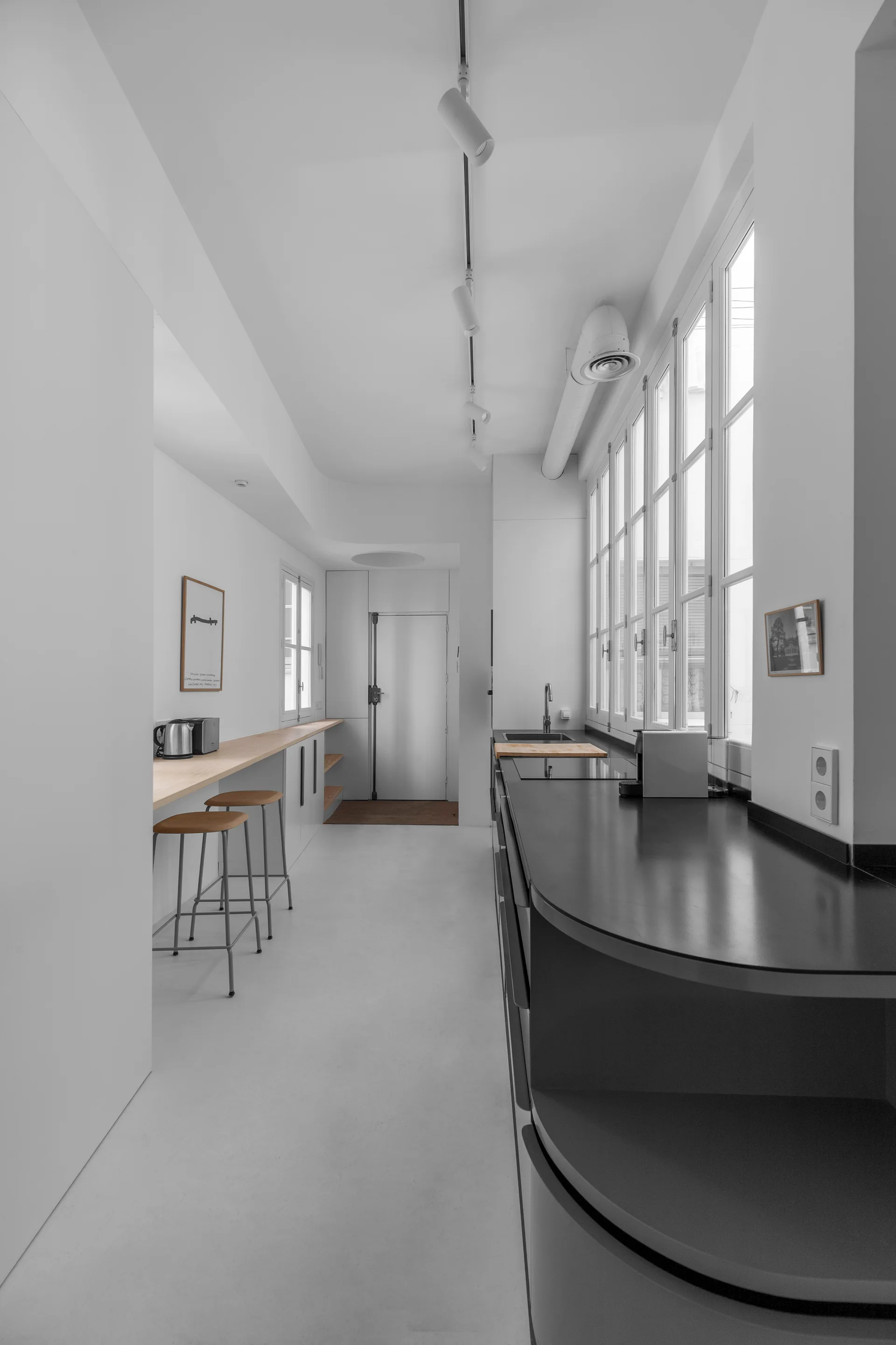 A minimalist galley kitchen featuring a long dark countertop under tall white windows opposite a light wood bar with metal stools.