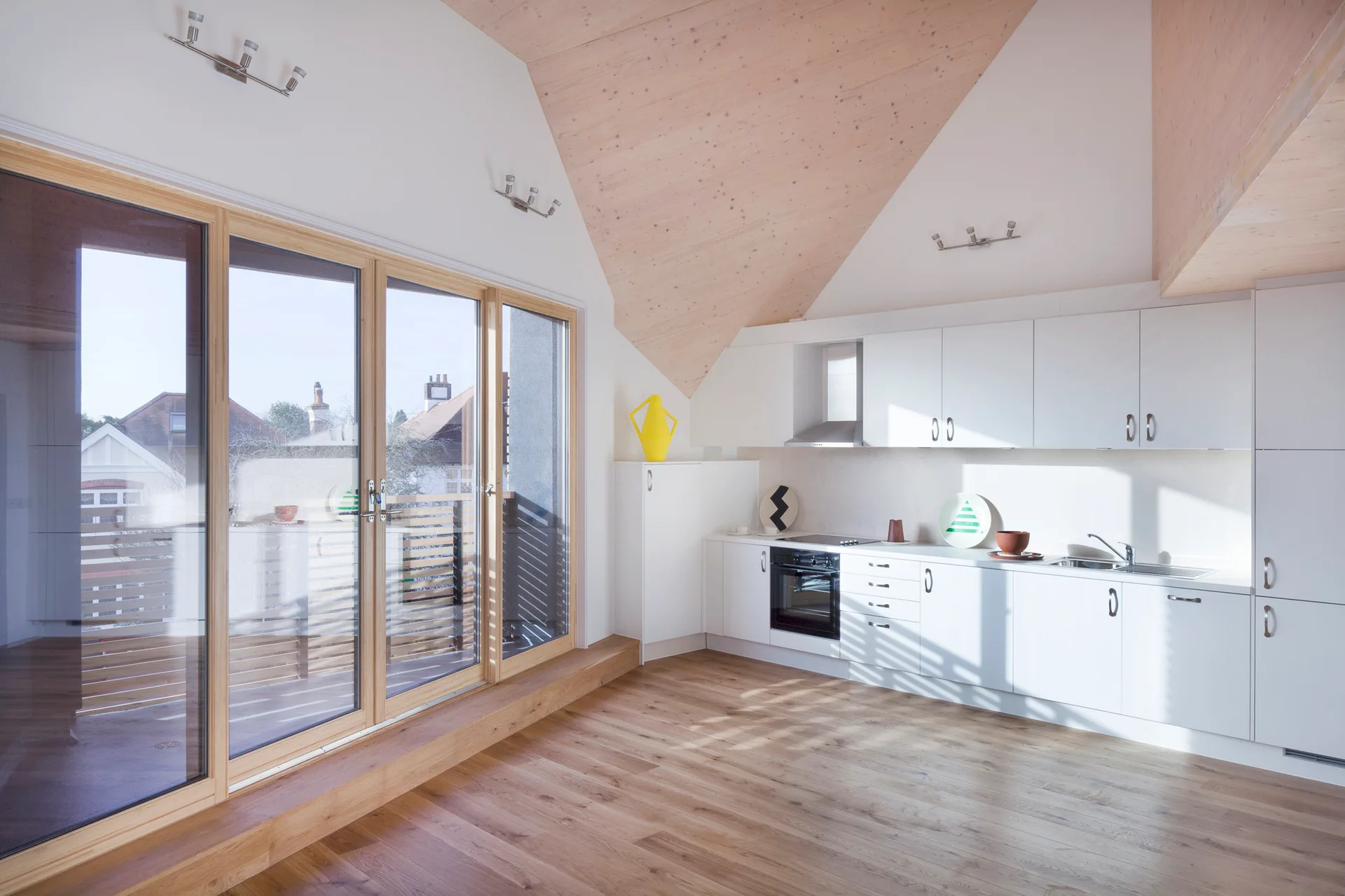 A minimalist white kitchen with flat-front cabinetry, light wood floors, a sloped wood ceiling, and large wooden-framed sliding glass doors.