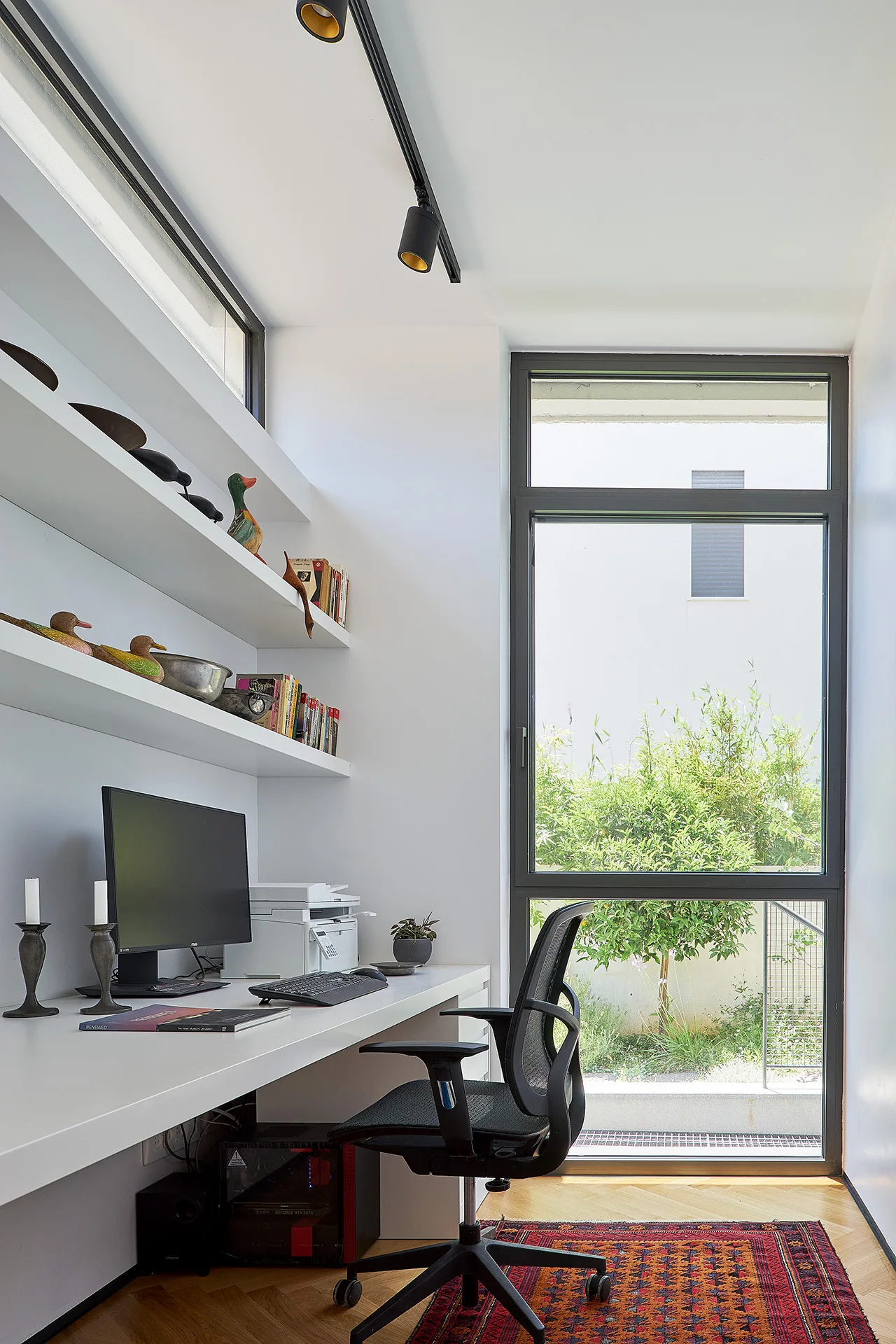 A modern home office featuring a white built-in desk, floating shelves, a black mesh chair, and a large window looking out onto a garden.
