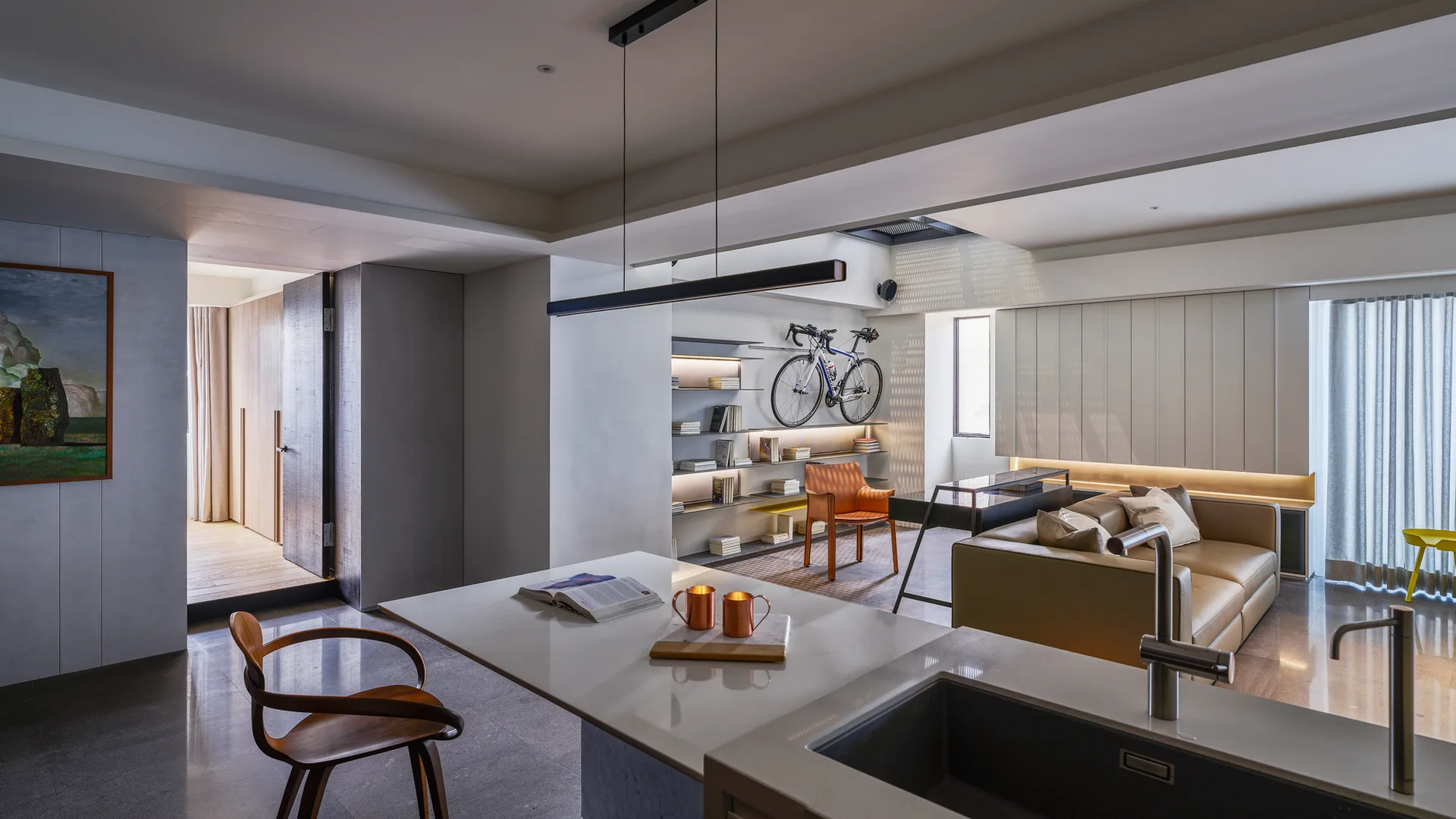 An open-concept living area and kitchen featuring a beige leather sofa, minimalist backlit shelving with a mounted bicycle, and a white kitchen island.