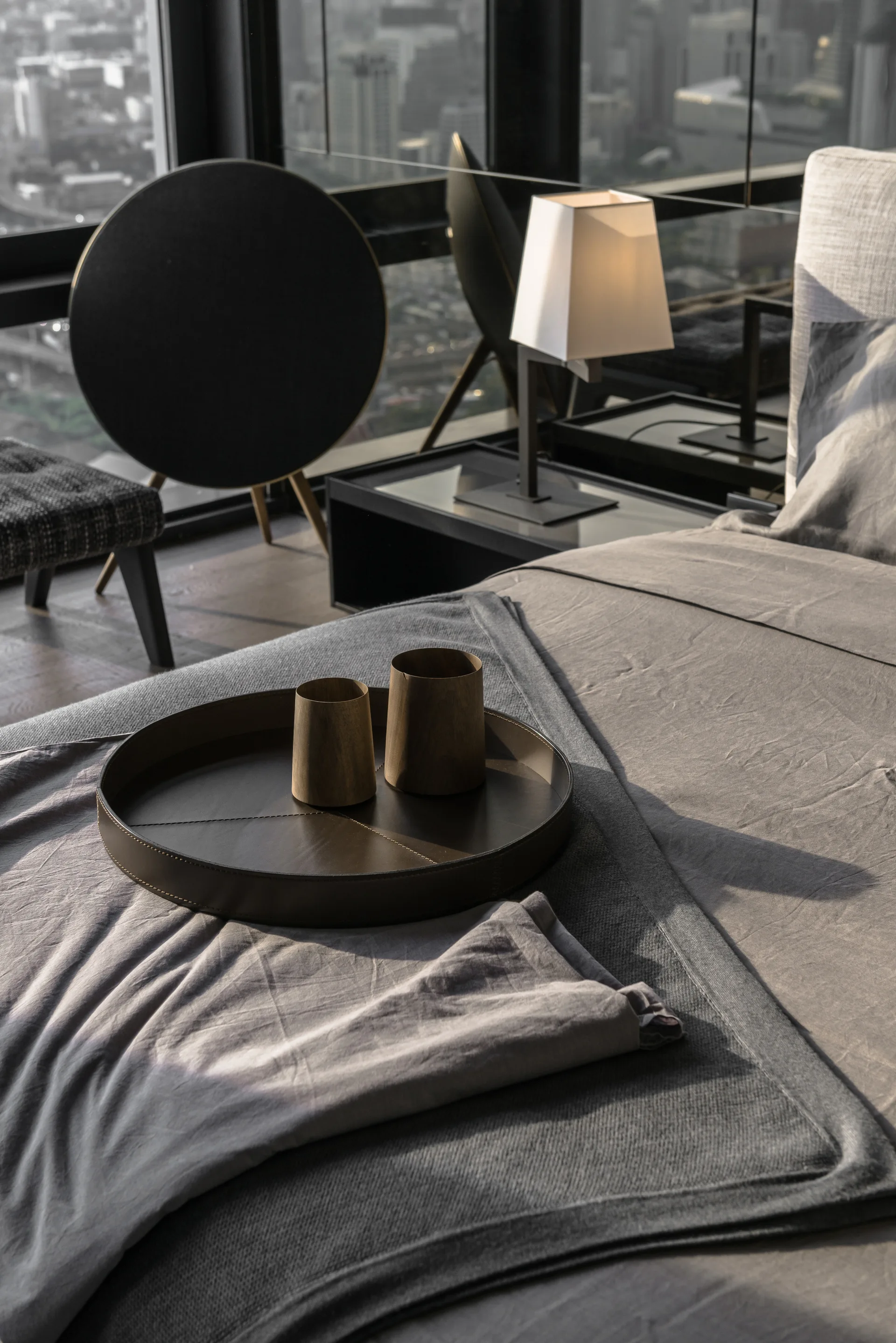 A close-up of a grey bed with a round leather tray holding wooden cups, set against a background of a nightstand lamp and high-rise window view.